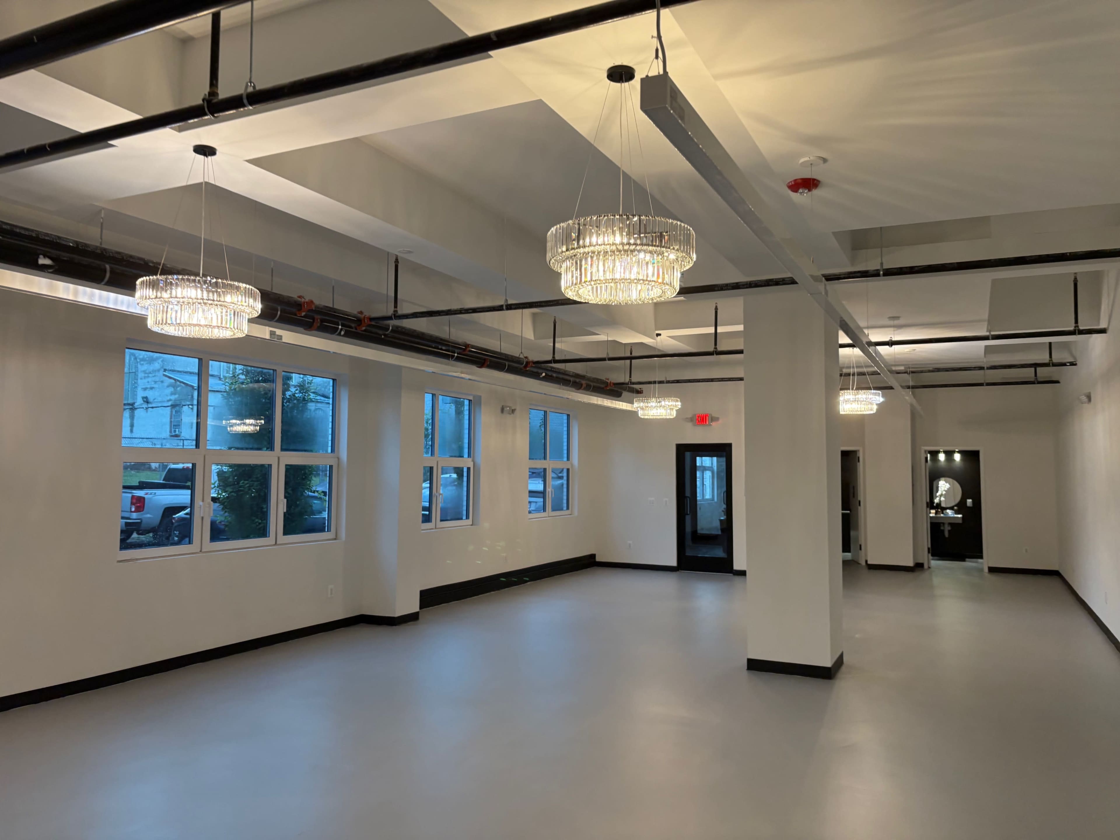 The image shows an empty, well-lit interior space with high ceilings, large windows, and modern light fixtures.