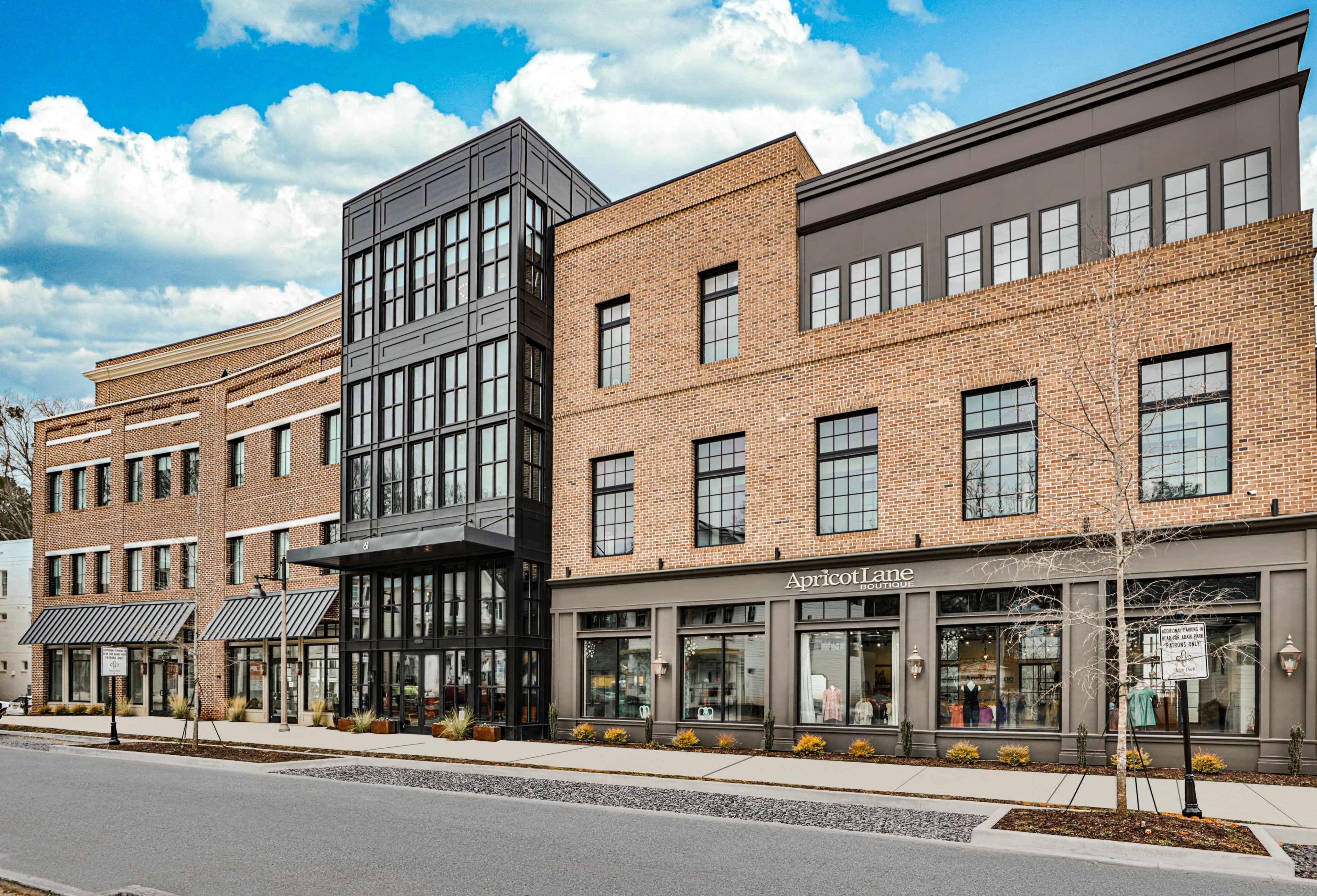 A modern multi-story brick building with large glass windows and retail spaces on the ground floor, located along a street lined with planters.