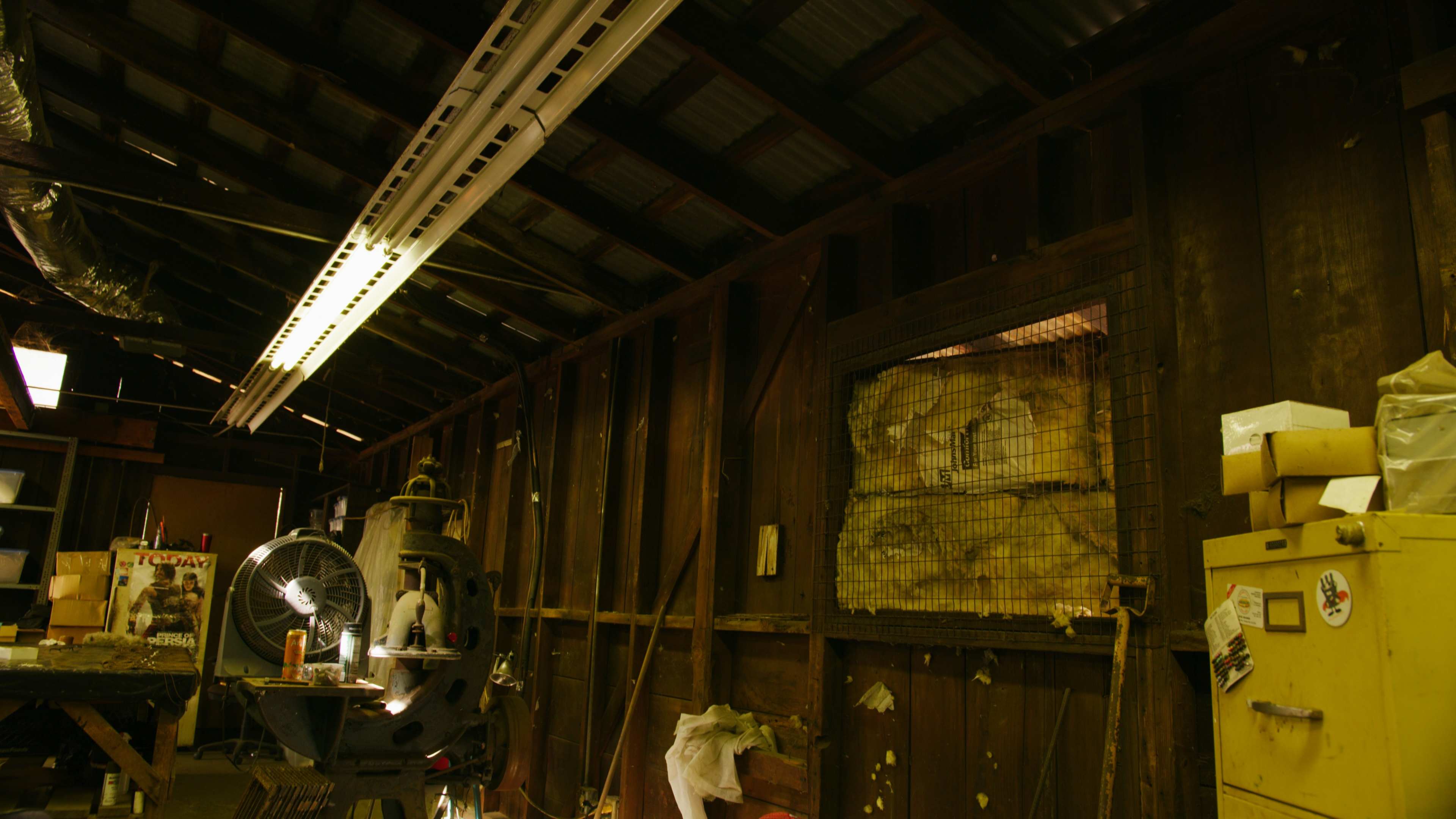 The interior of a dimly lit workshop with wooden walls, tools, and a fan, featuring a window covered by a wire mesh.