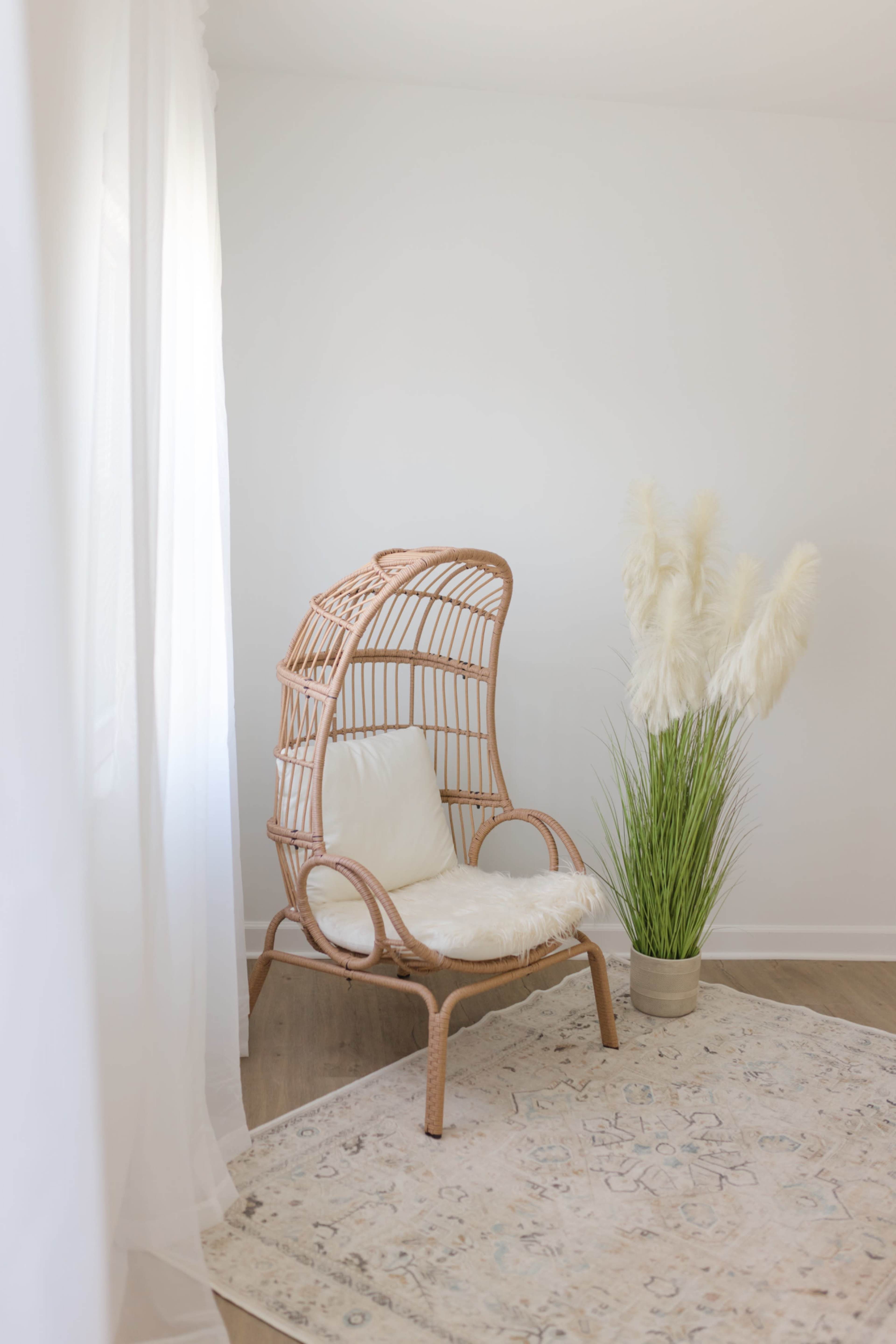 A rattan chair with a high back sits beside a potted grass plant, all set against a light, minimalistic interior.