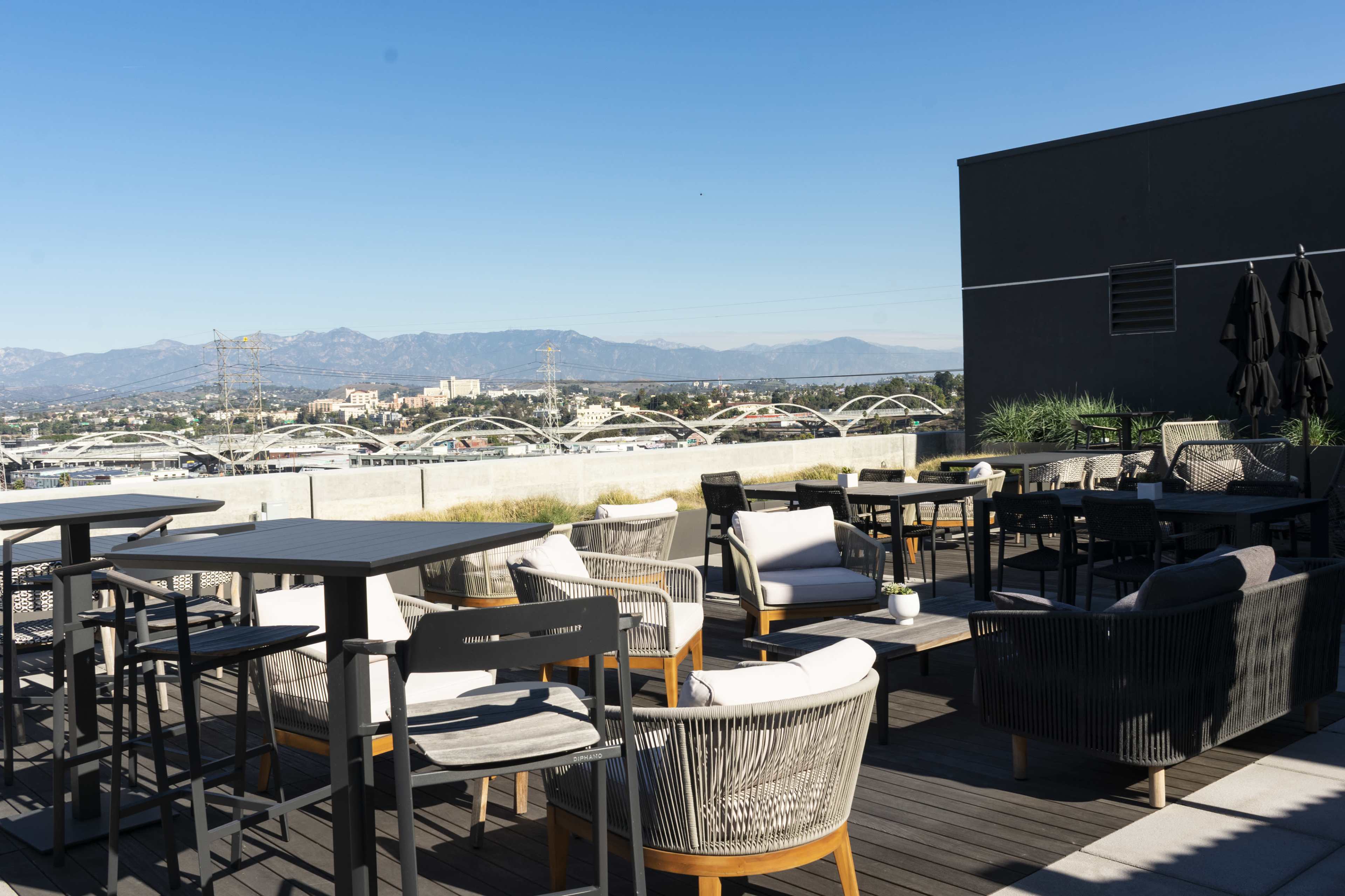 A rooftop terrace filled with modern seating arrangements and tables, overlooking a scenic view of mountains and city buildings.