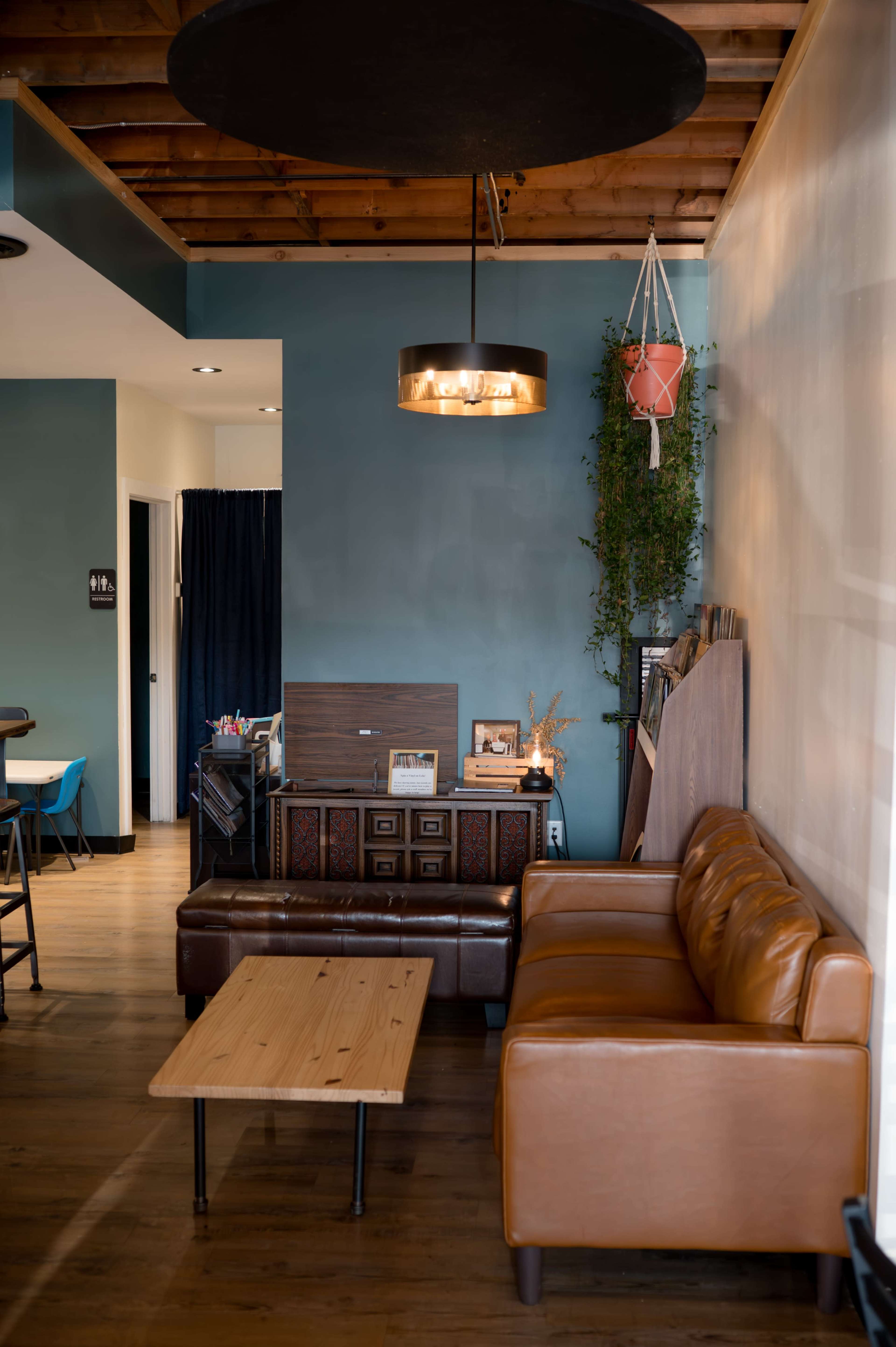 The image shows a cozy café interior featuring a brown leather couch, a wooden coffee table, and a decorative wall with plants and artwork.