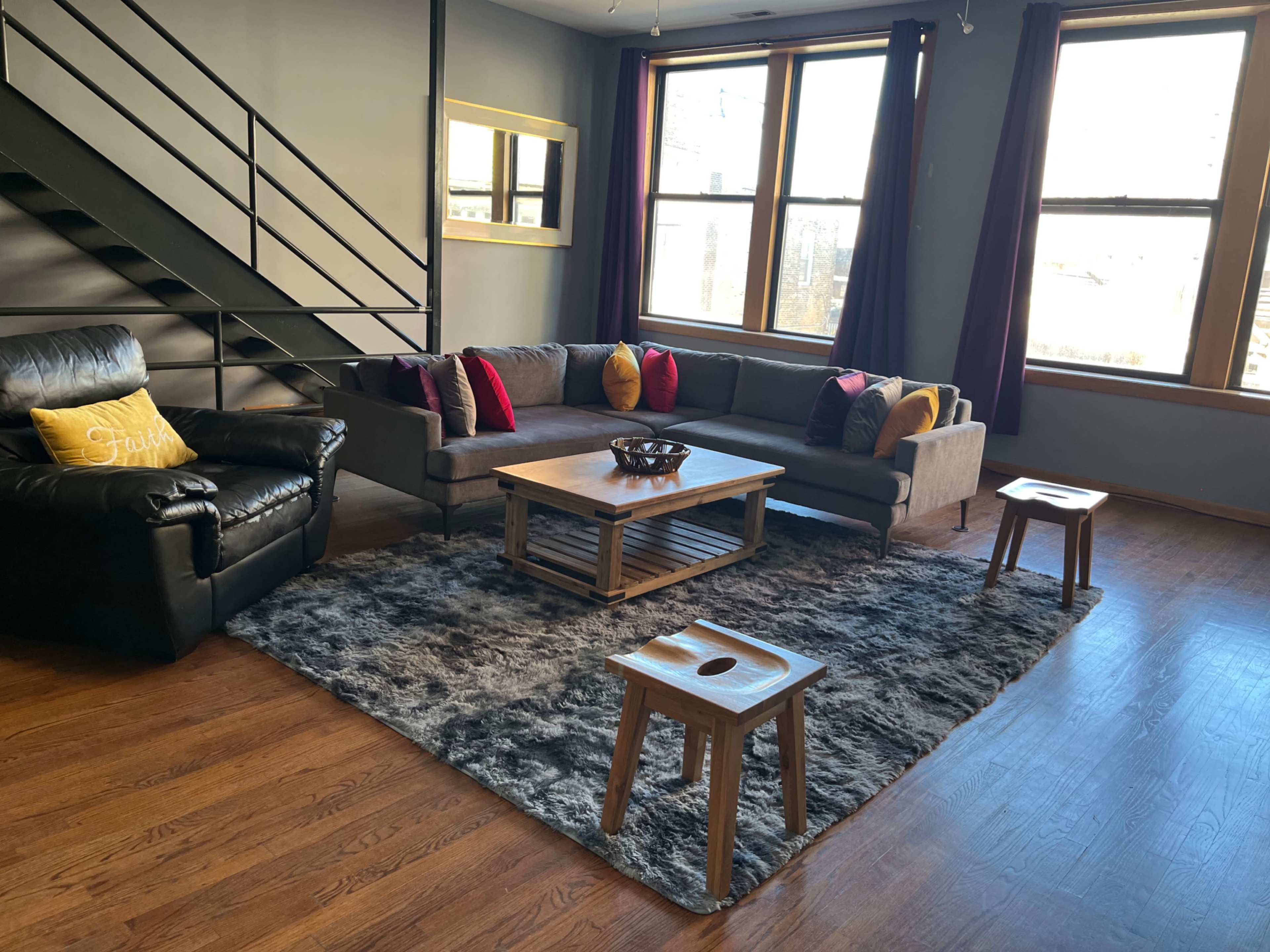 The image shows a modern living room with a sectional sofa, a coffee table, and two small side tables, all set on a large area rug, with large windows letting in natural light.