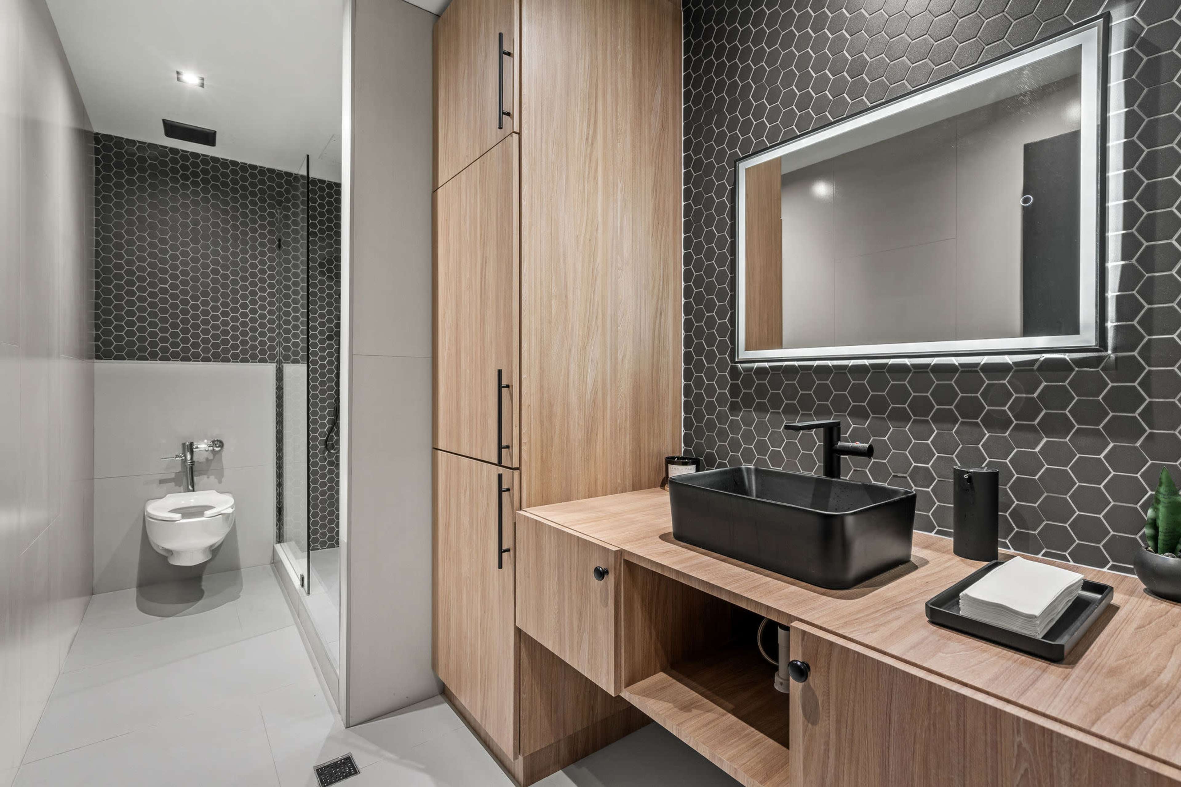 The bathroom features a modern design with a hexagonal tile wall, wooden cabinetry, a black vessel sink, and a wall-mounted mirror.