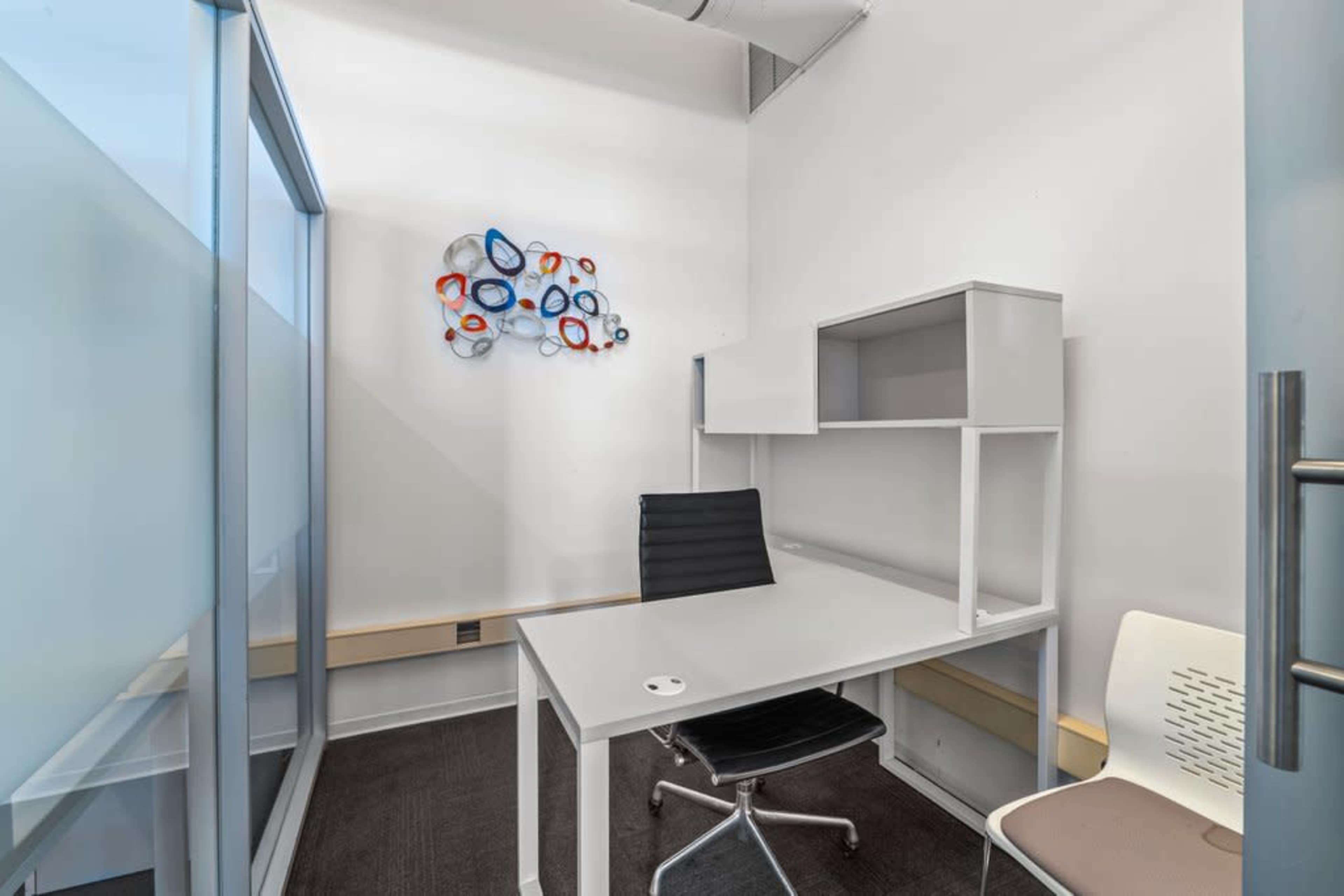 The image shows a modern, minimalistic office space with a white desk, a black office chair, and a decorative wall piece.