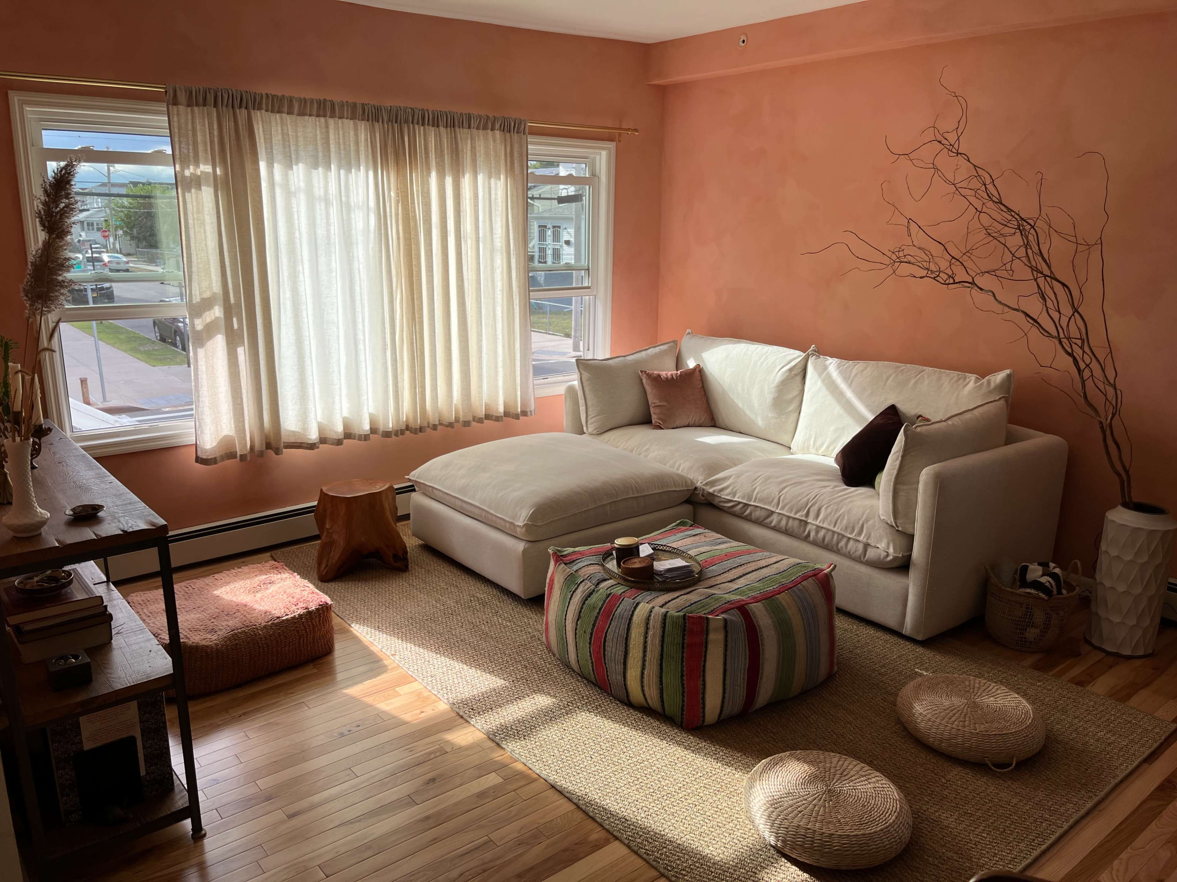 A cozy living room with a large white sectional sofa, a wooden coffee table, woven decor, and a large window with sheer curtains letting in natural light.