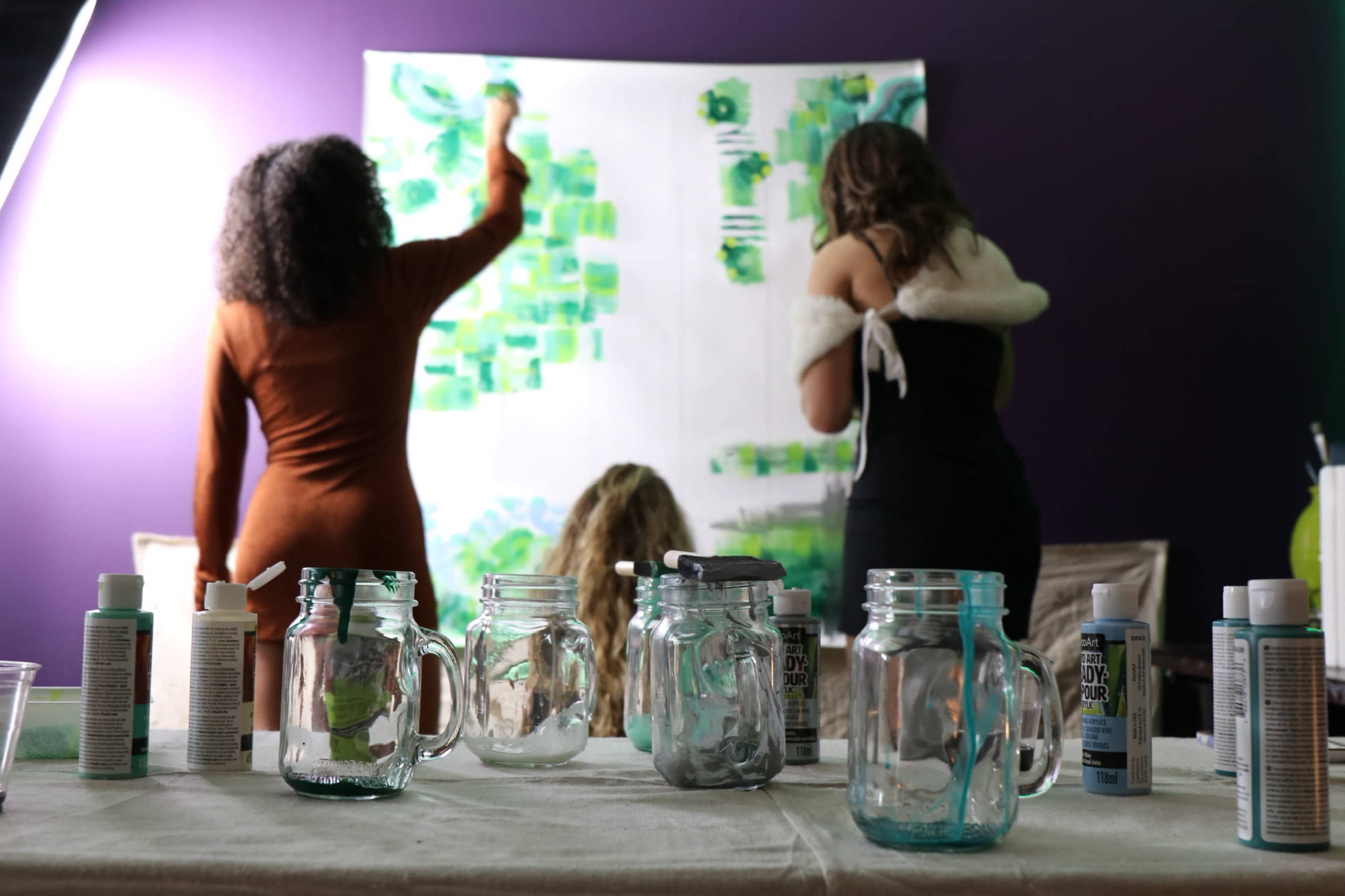 Three women are painting a large canvas on a purple wall, with glass jars and paint supplies arranged on a table in the foreground.