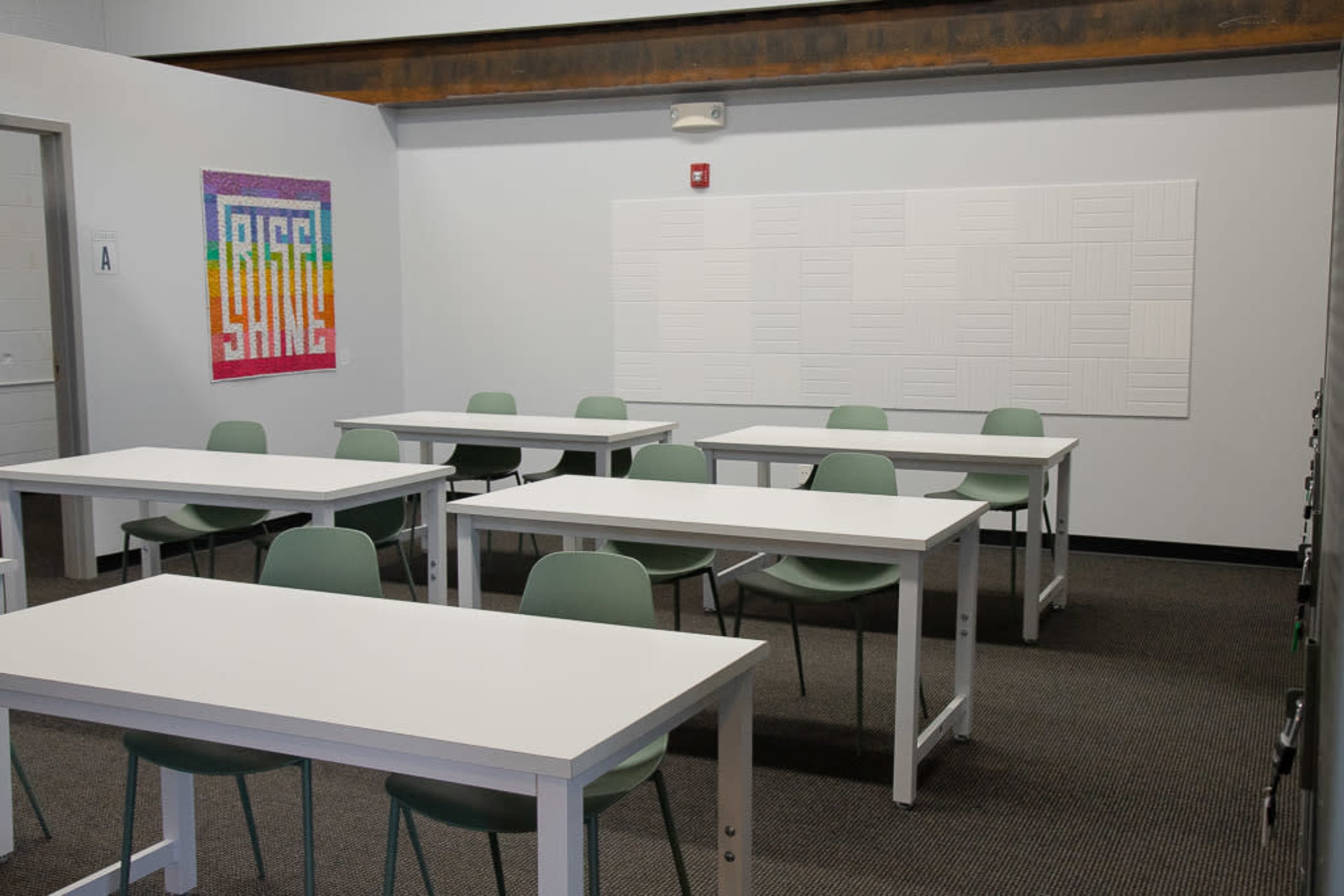An empty classroom with several white tables and green chairs arranged neatly, along with a colorful "RISE" mural on one wall.