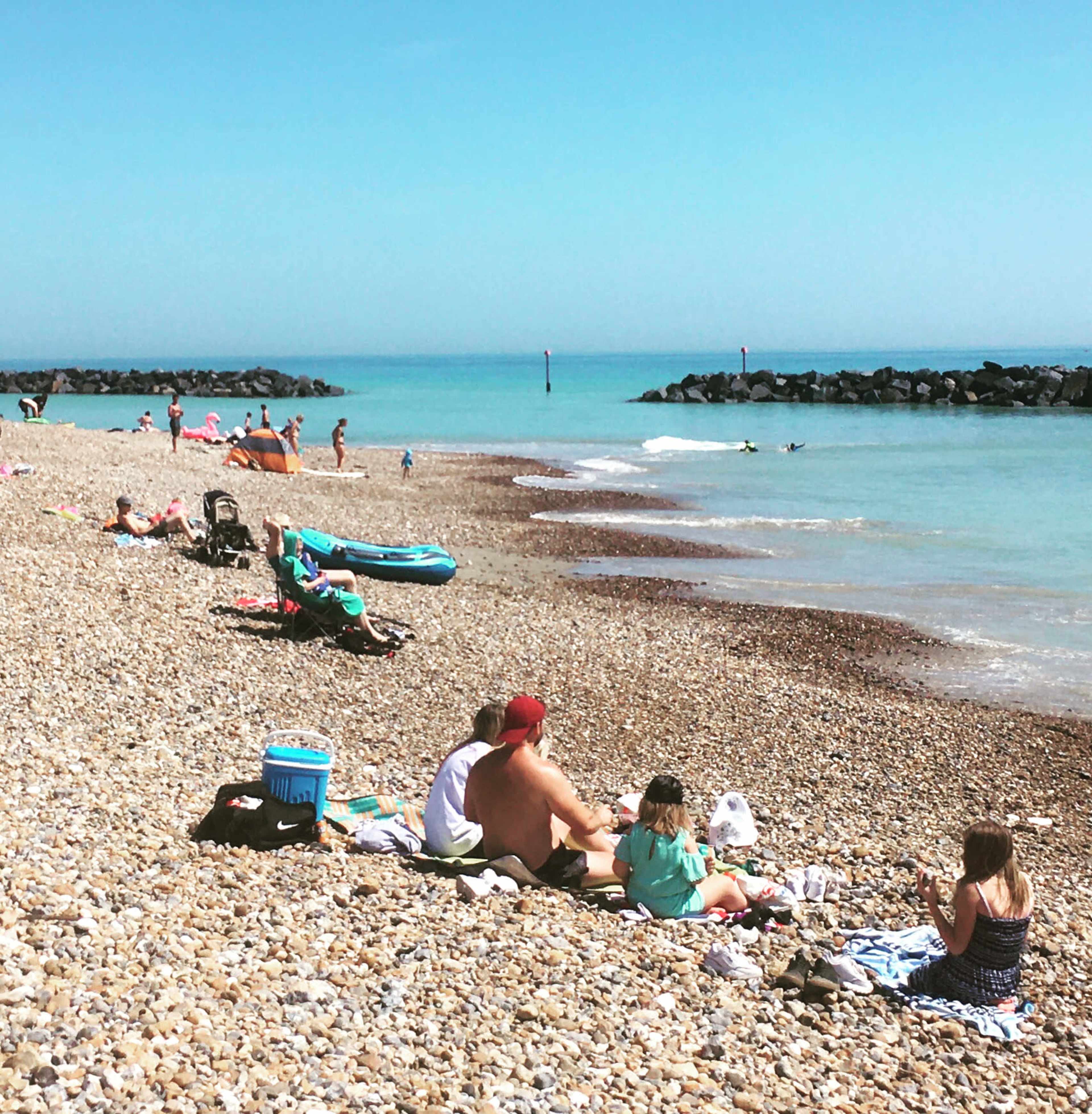 People relax on a pebbled beach under a clear blue sky, with some swimming in the water and others lounging on towels.