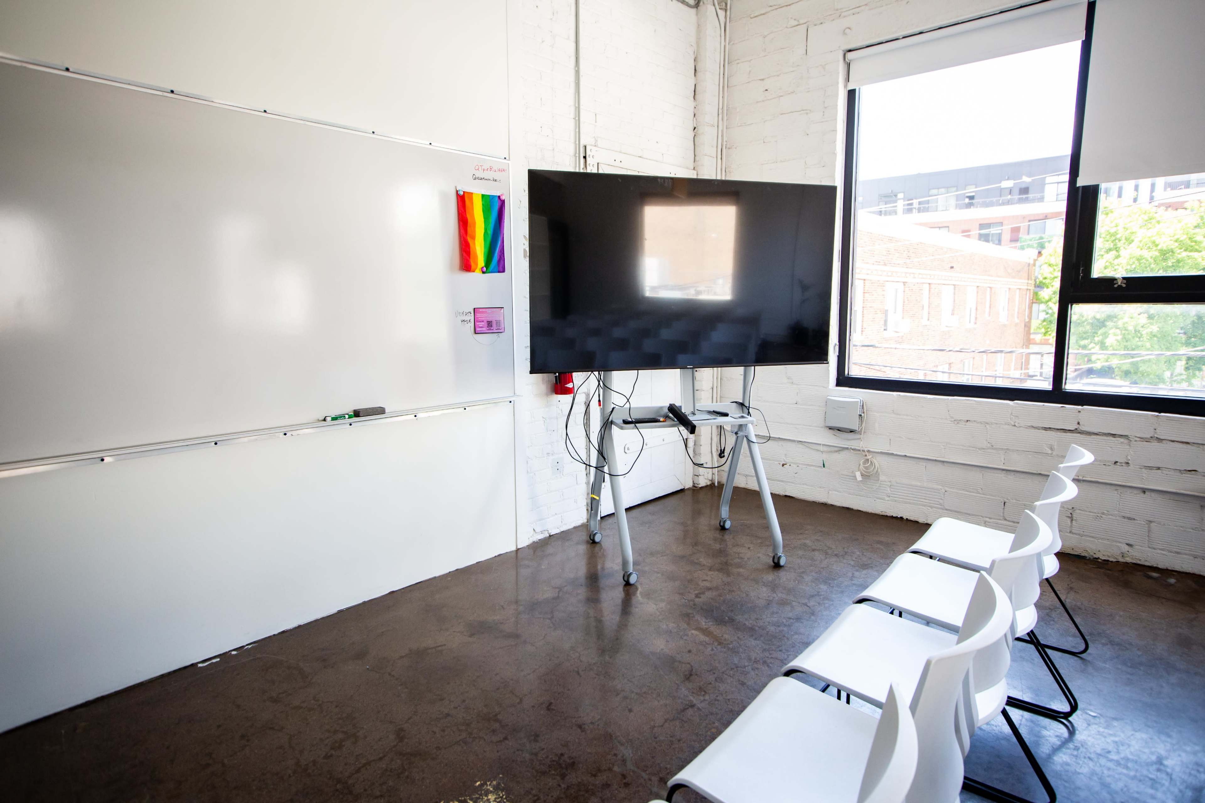 A modern classroom features a large screen mounted on a stand, a whiteboard on the wall, and several empty white chairs arranged in front.