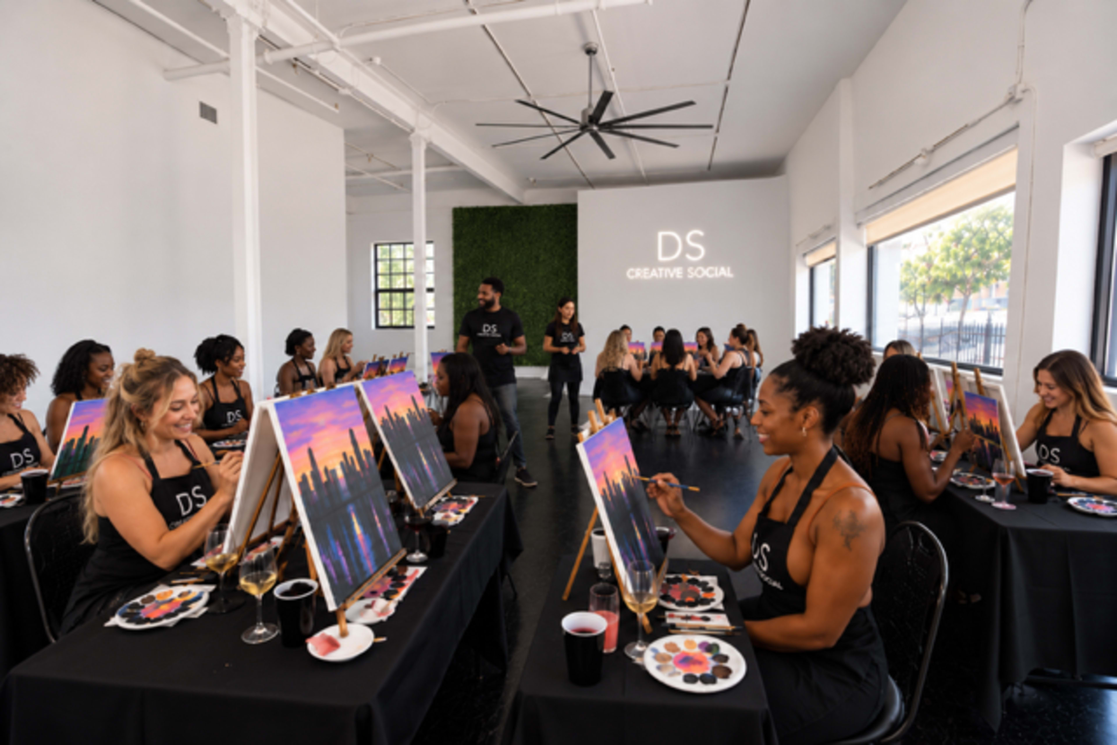 A group of people sitting at easels in a brightly lit room are participating in a painting class, with a city skyline scene displayed on their canvases.