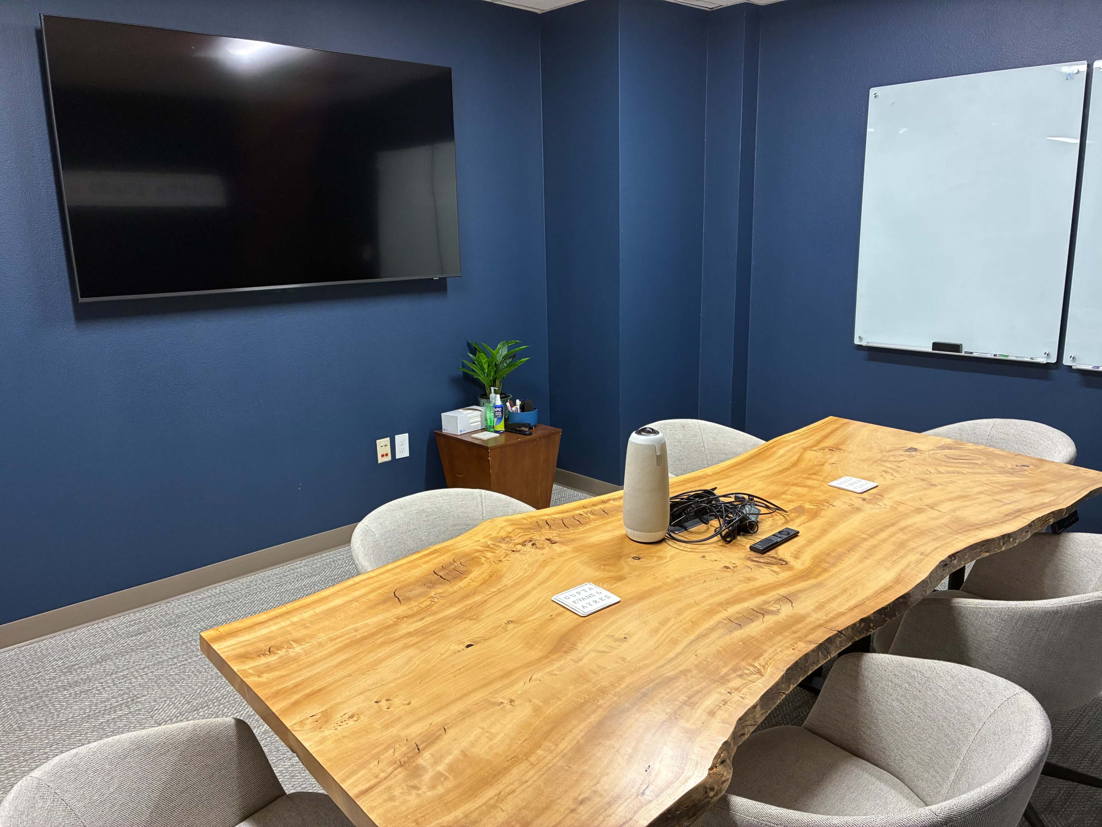 The image shows a modern conference room with a wooden table, six light-colored chairs, a large flat-screen TV on the wall, a plant on a side table, and a whiteboard.