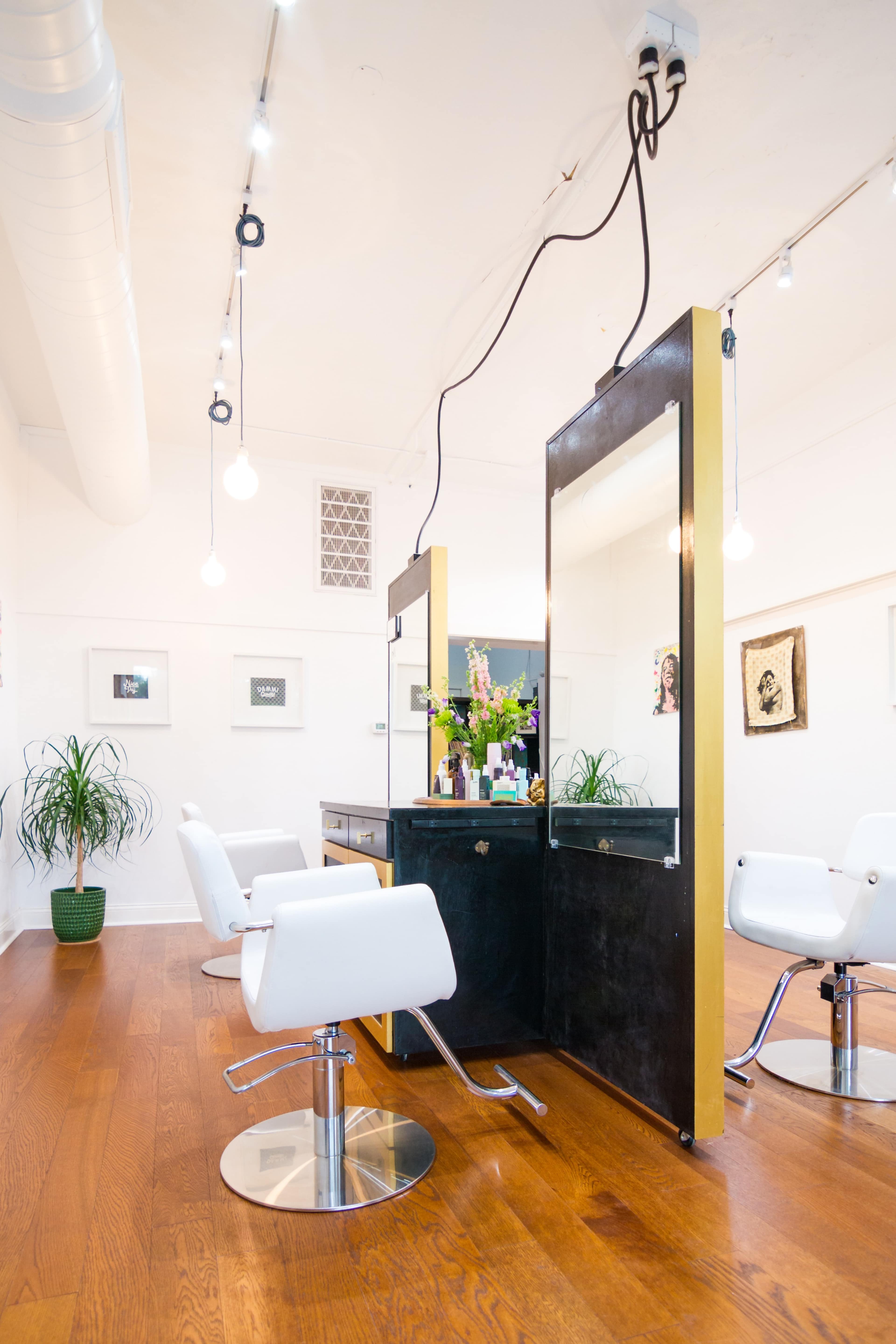 A modern hair salon interior featuring two styling stations with mirrors, sleek white chairs, and wooden flooring, illuminated by pendant lights.