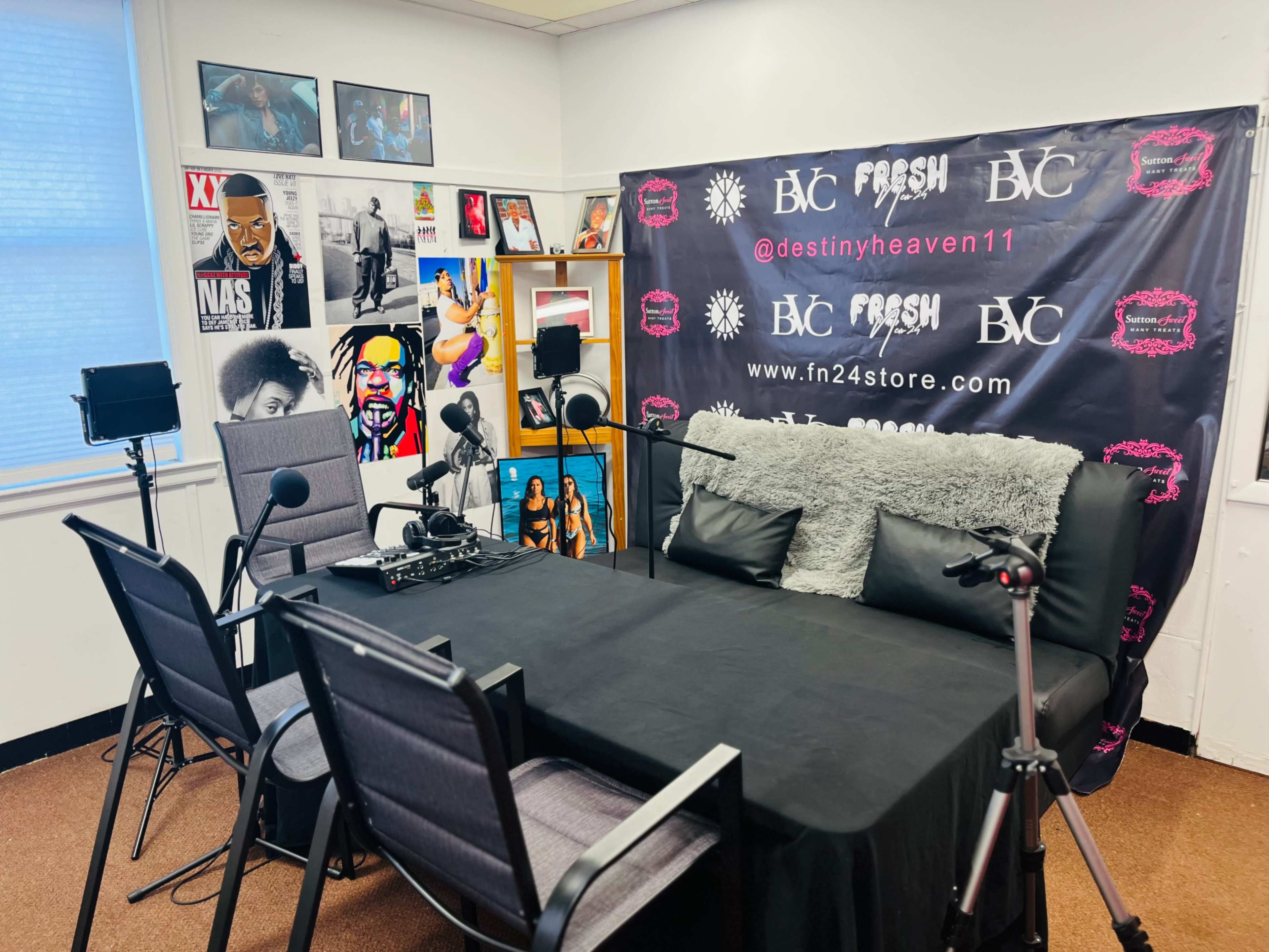 A recording studio setup with a table, microphones, and chairs, surrounded by framed posters on the walls.