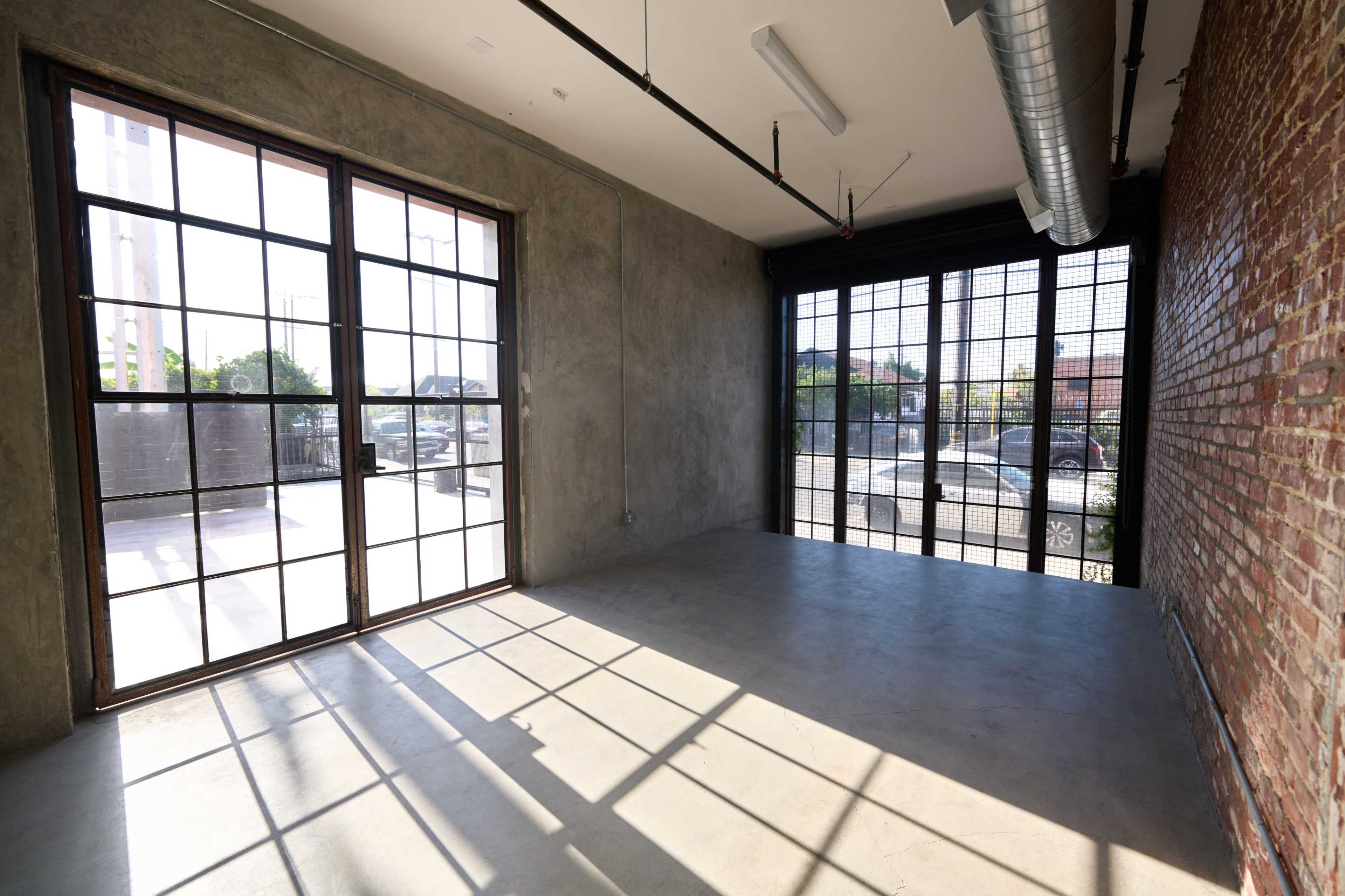 The image shows a minimalist industrial-style room with large windows, concrete flooring, and exposed brick walls.