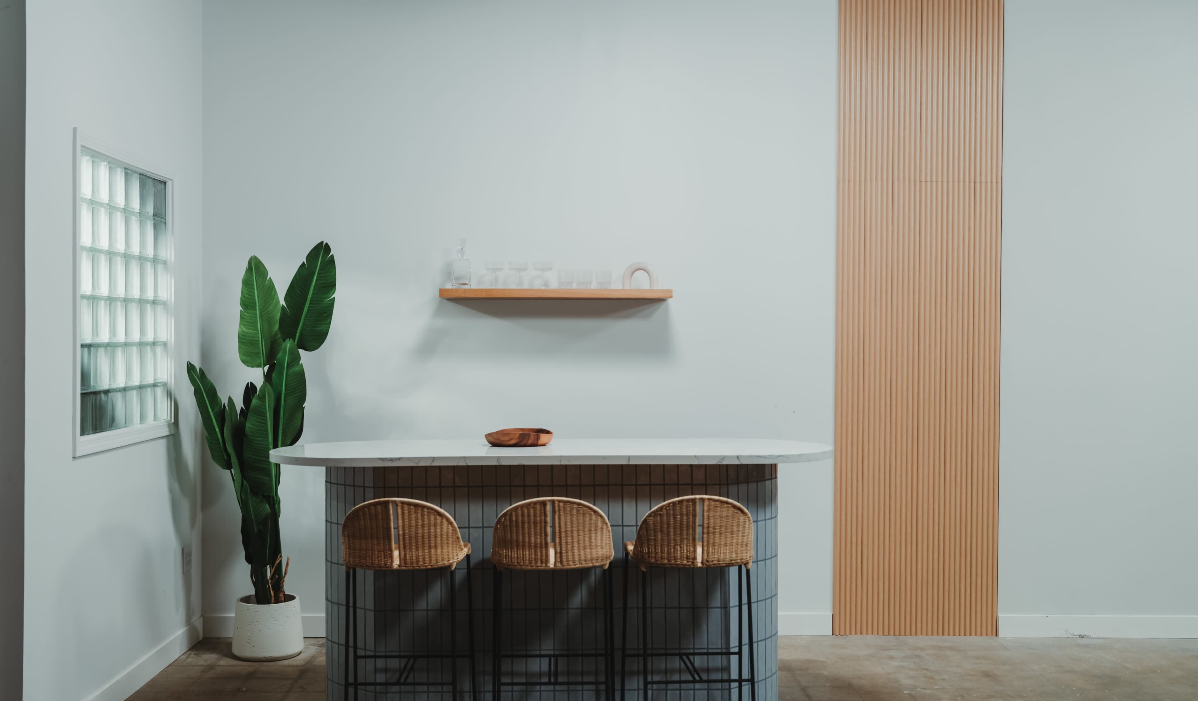 A modern kitchen features a bar-style counter with three woven chairs, a green plant in a pot, and a wooden shelf mounted on a light wall.