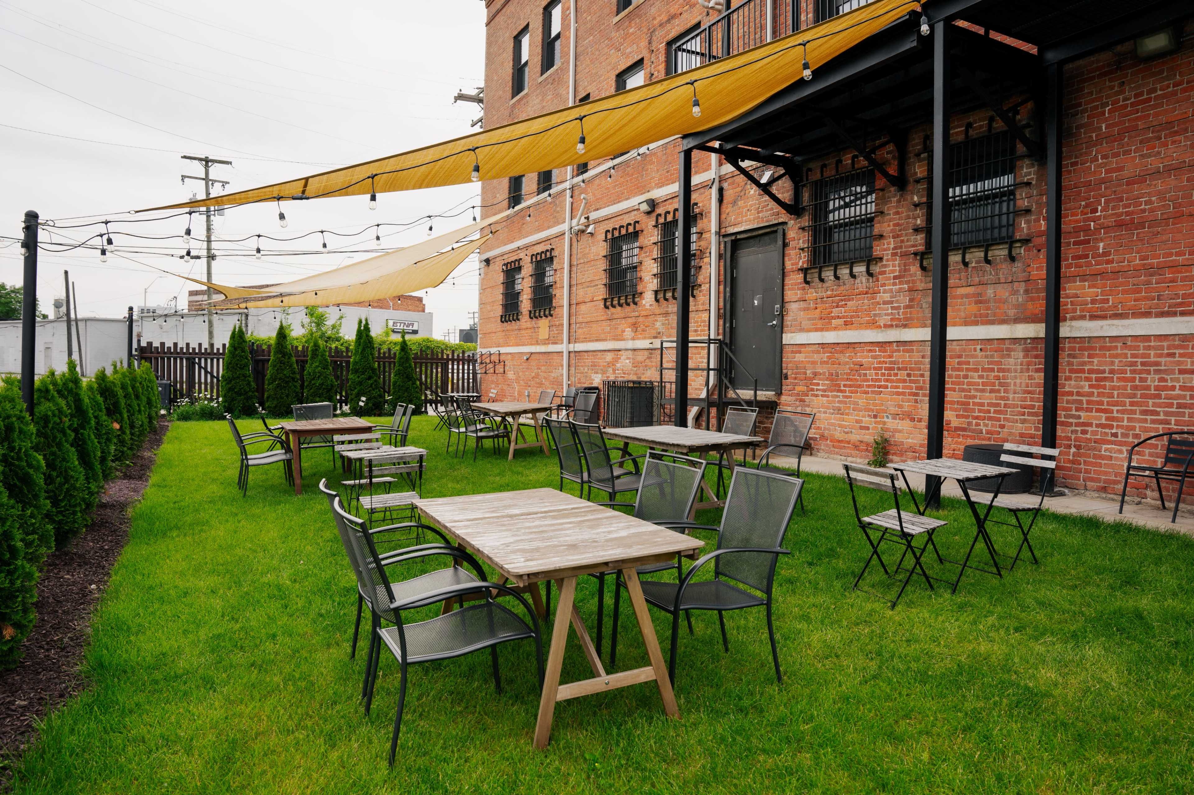 An outdoor seating area with several wooden and metal tables and chairs arranged on a grassy lawn beside a brick building.
