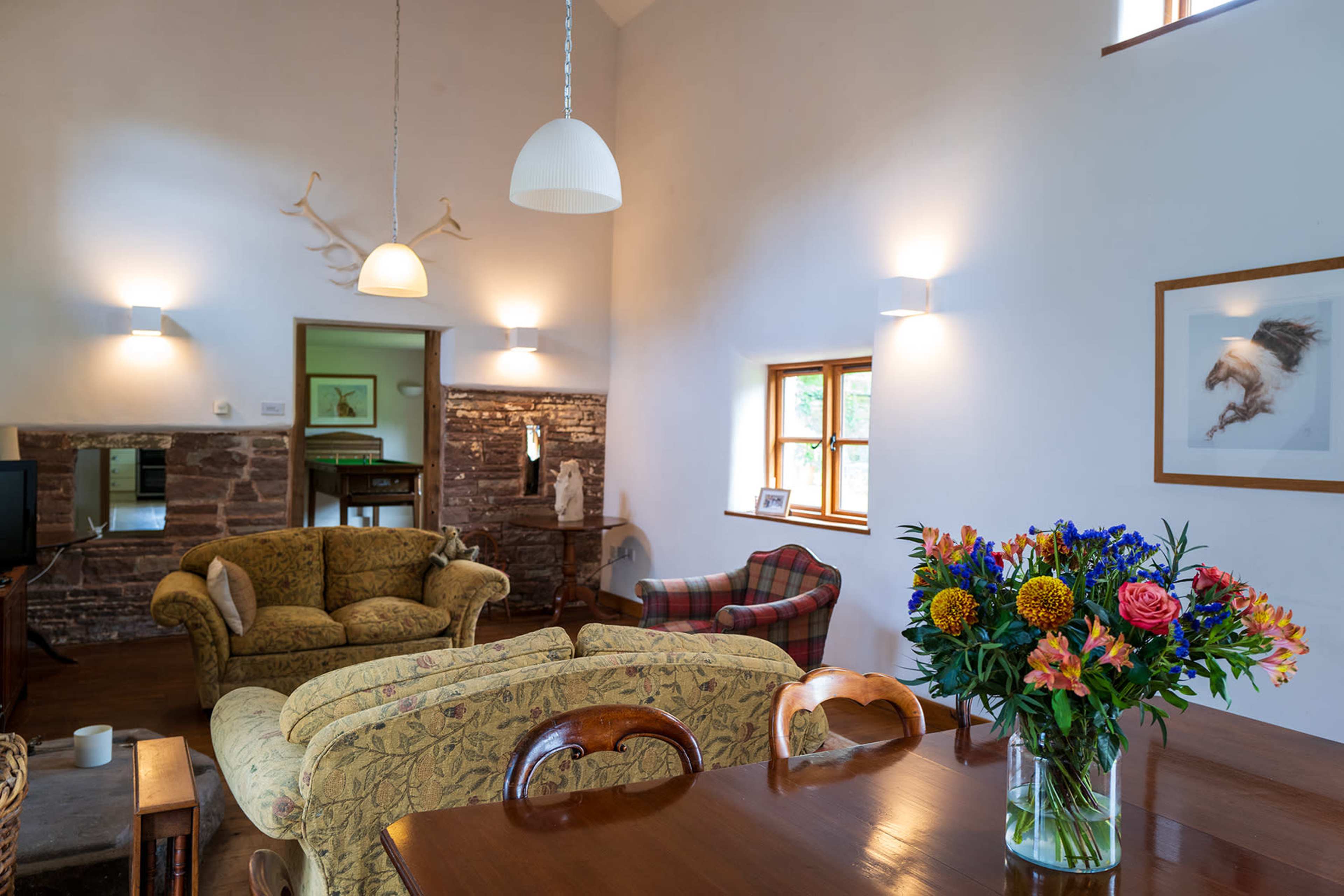 A cozy living area with a green floral sofa, a plaid armchair, a wooden dining table, and a vase of colorful flowers on the table, illuminated by wall sconces and pendant lights.