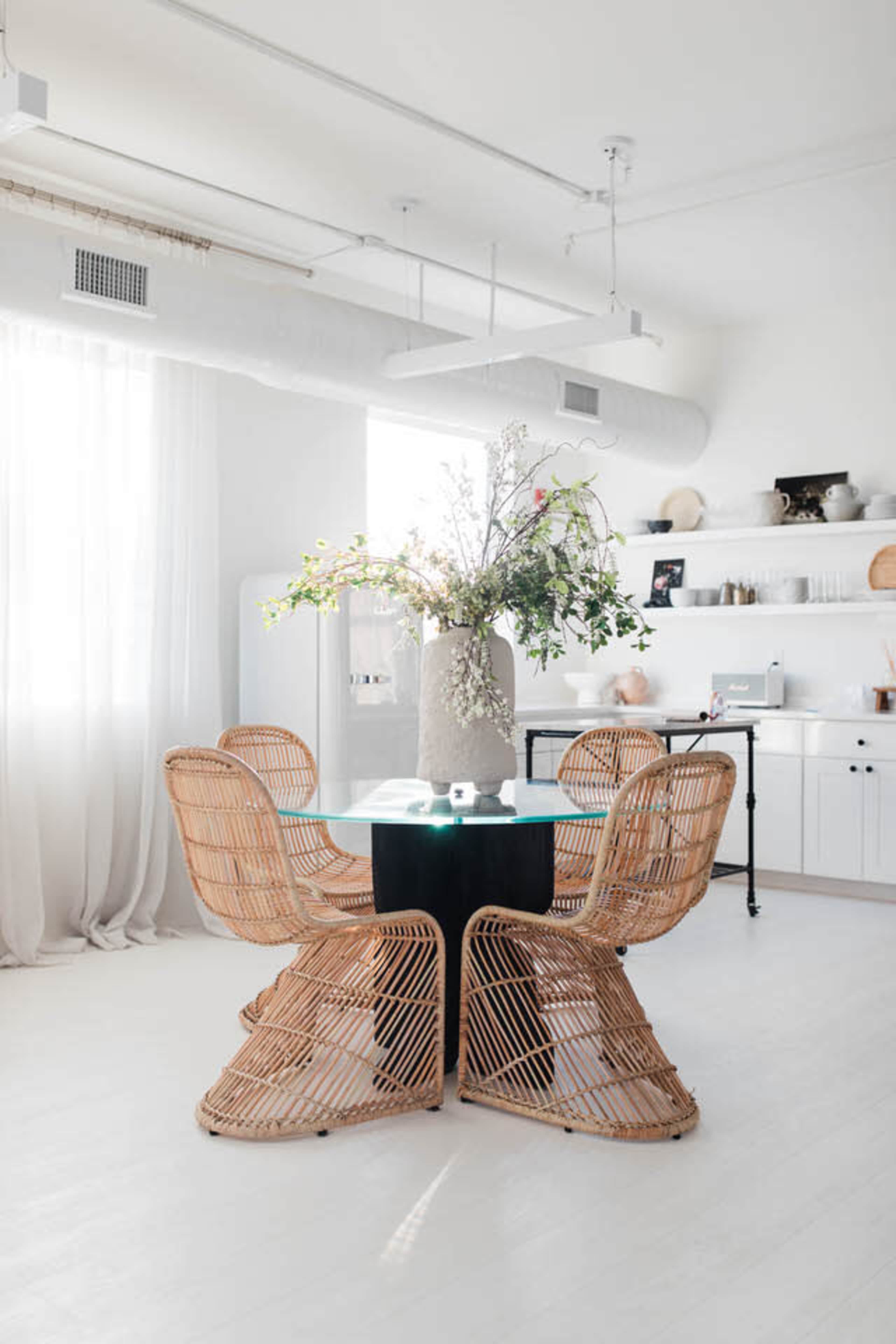 A round table with a glass top and a large vase filled with greenery is surrounded by four rattan chairs in a bright, minimalist dining area.