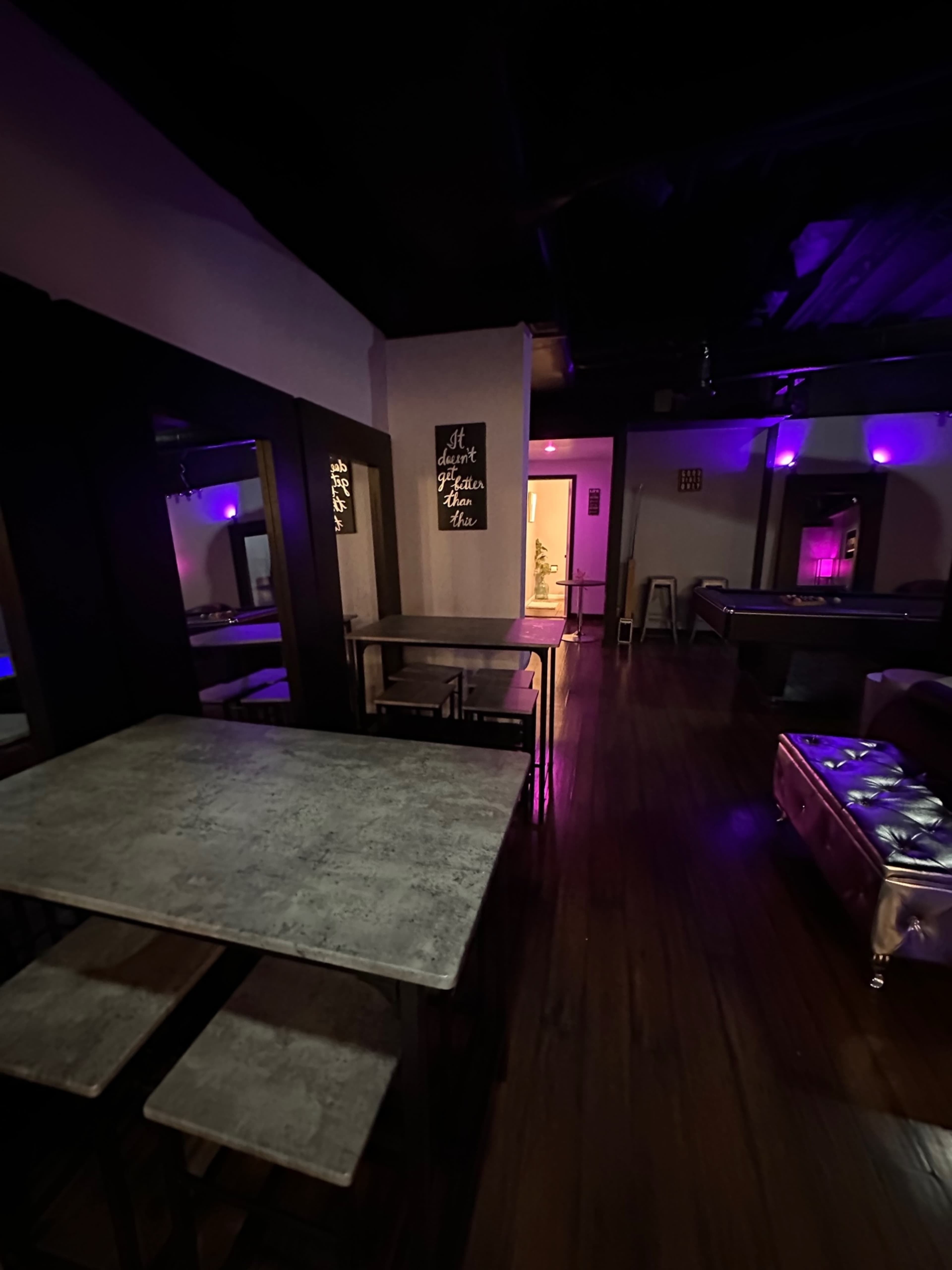 A dimly lit lounge area with purple lighting, featuring a wooden floor, a metallic sofa, and tables arranged near the walls.