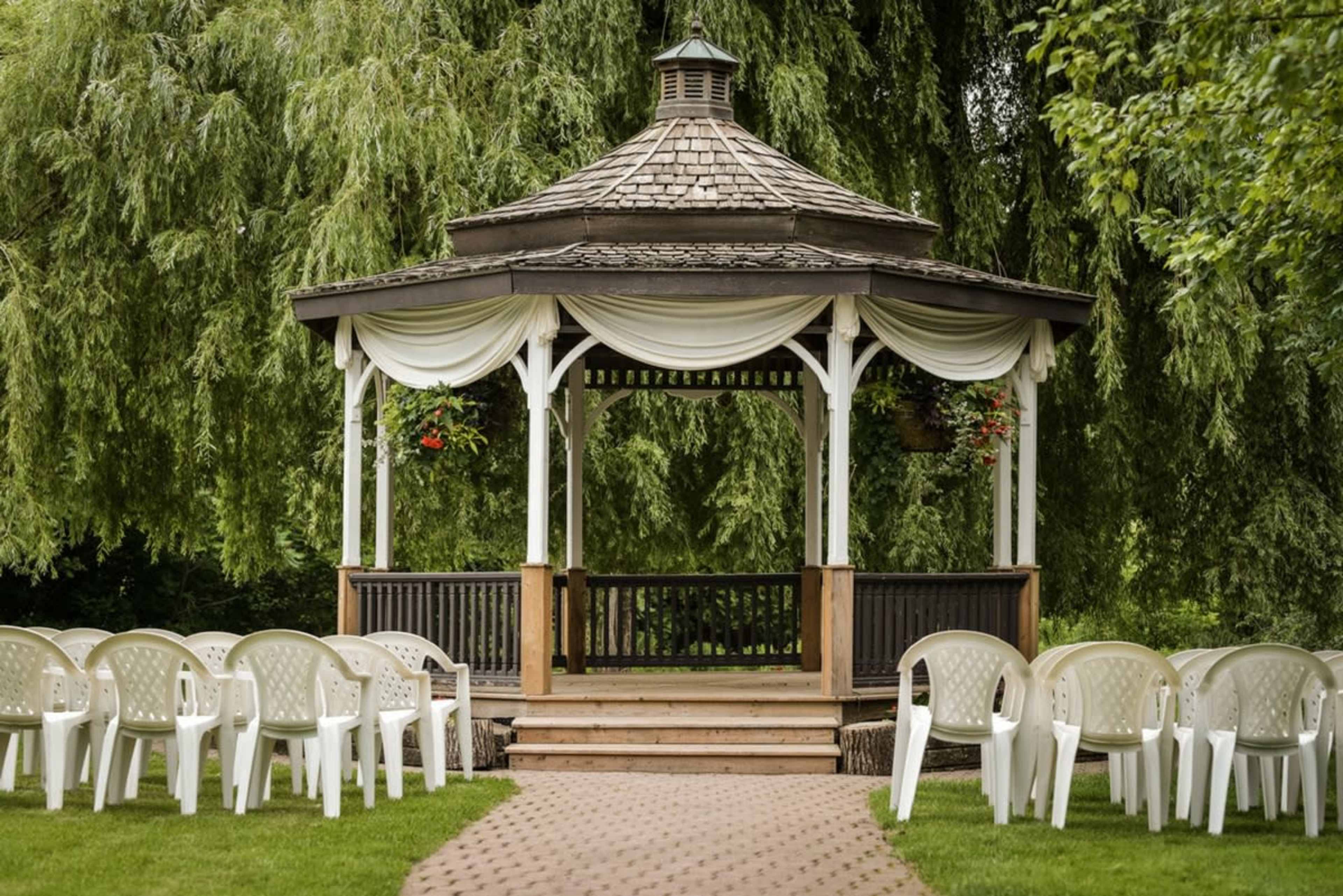 A gazebo with draped fabric stands at the end of a path lined with chairs in a green outdoor setting.