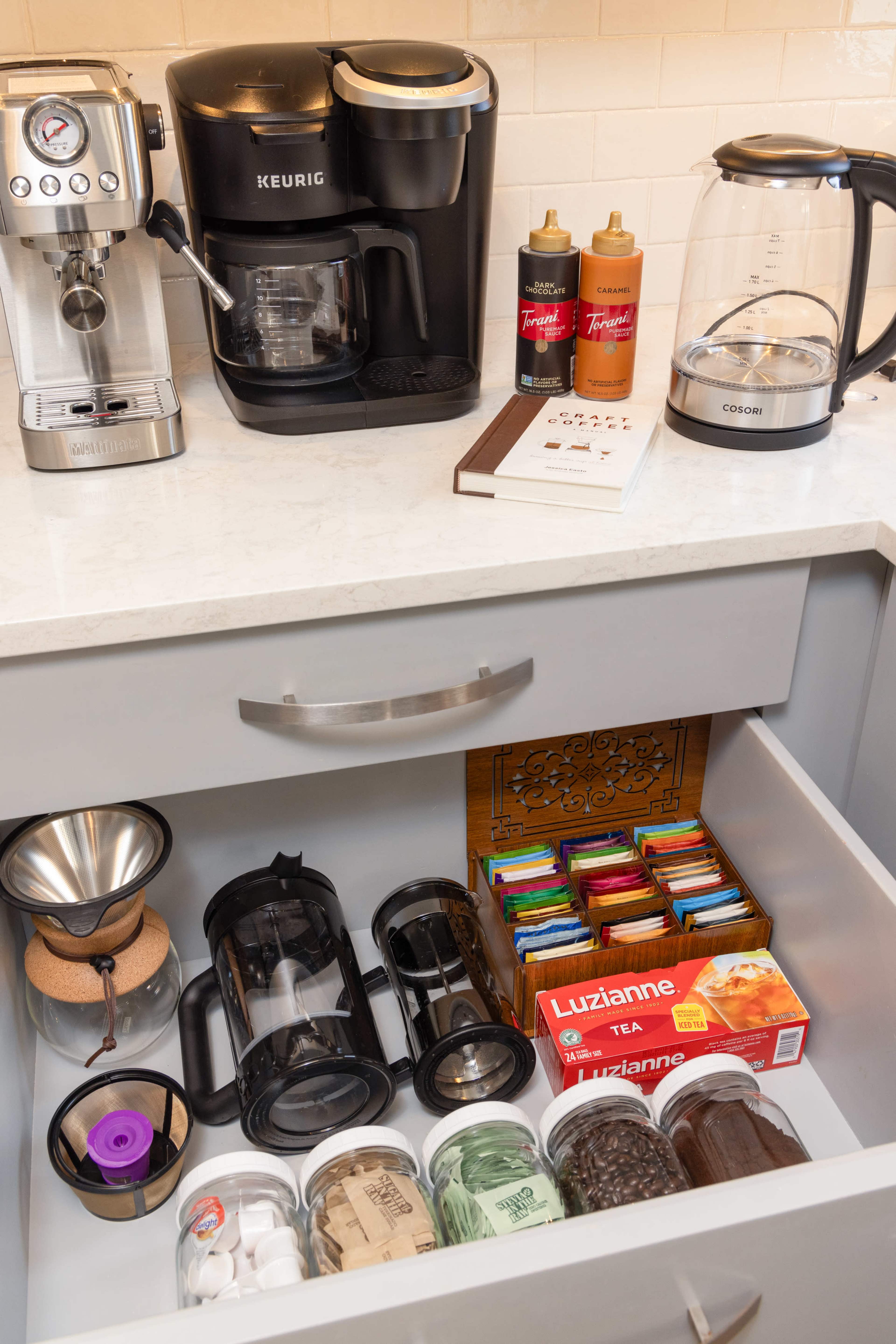 A kitchen countertop with a coffee machine, a kettle, and a drawer containing various coffee and tea supplies, including a French press and assorted tea bags.
