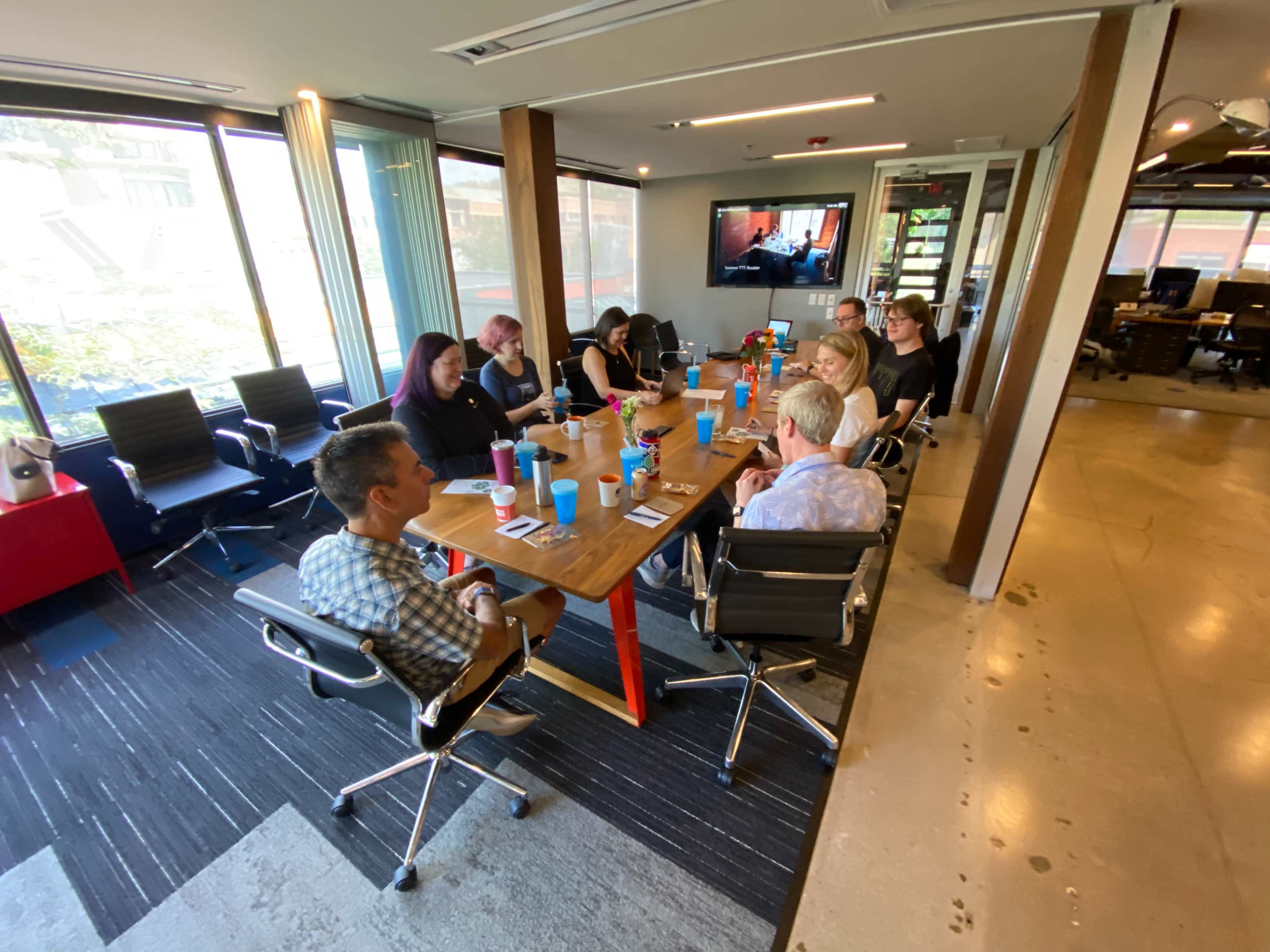 A group of eight people sits around a wooden table in a modern office, with a television displaying a presentation in the background.