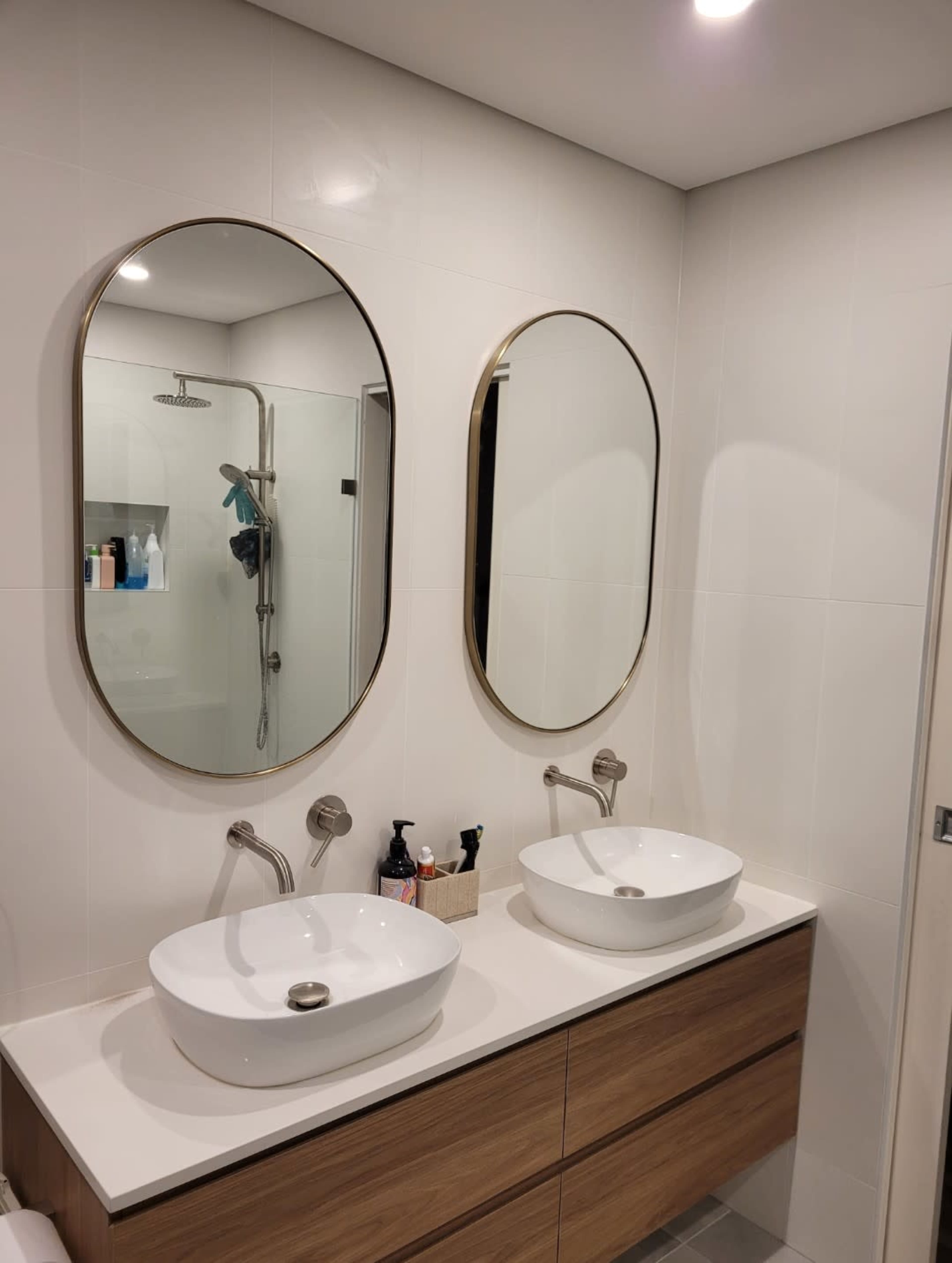 The image shows a modern bathroom with two oval mirrors above separate sinks on a wooden vanity, and a shower area visible in the background.
