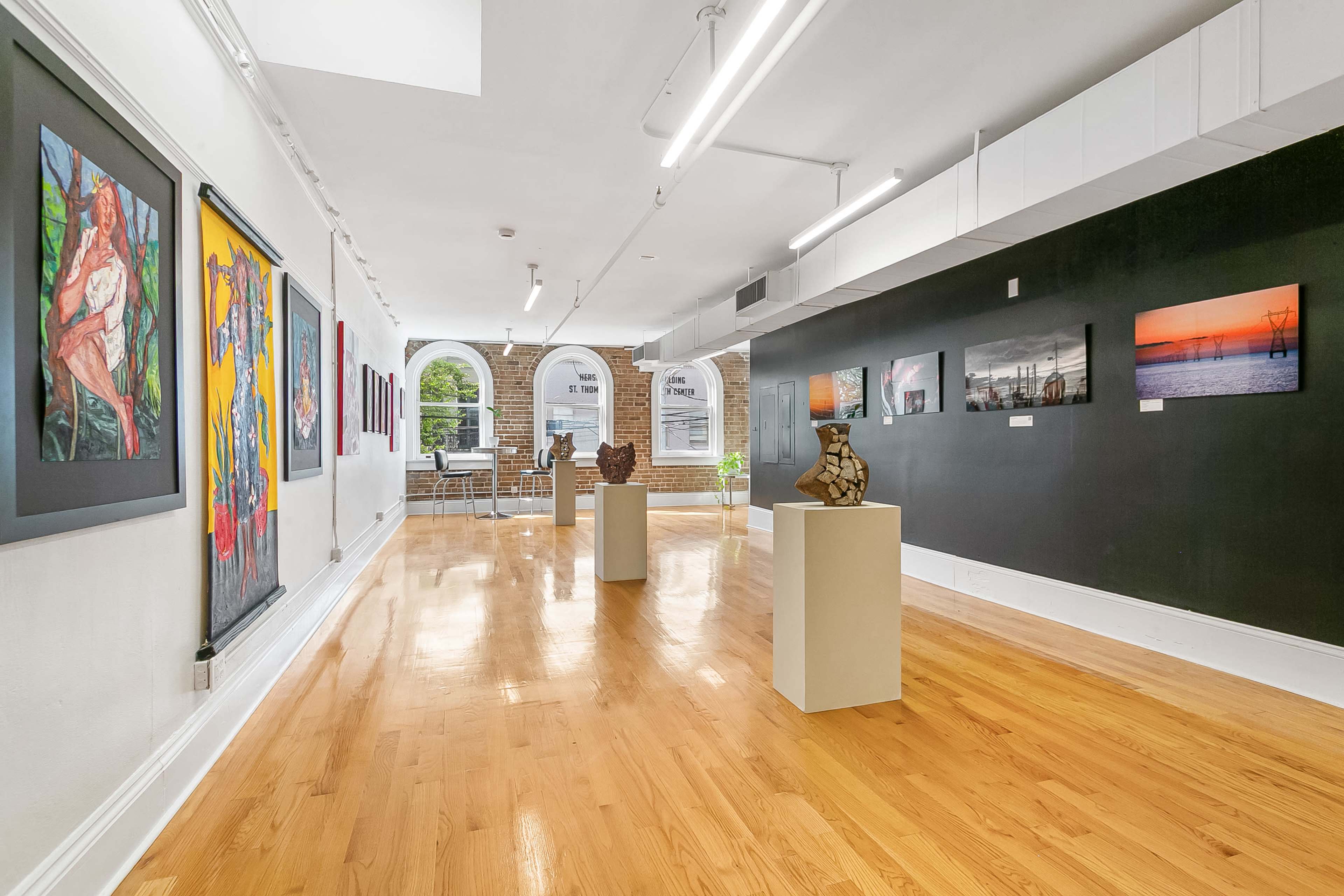 The image shows an art gallery with paintings and sculptures displayed along the walls and on pedestals, featuring a polished wooden floor and ample natural light.