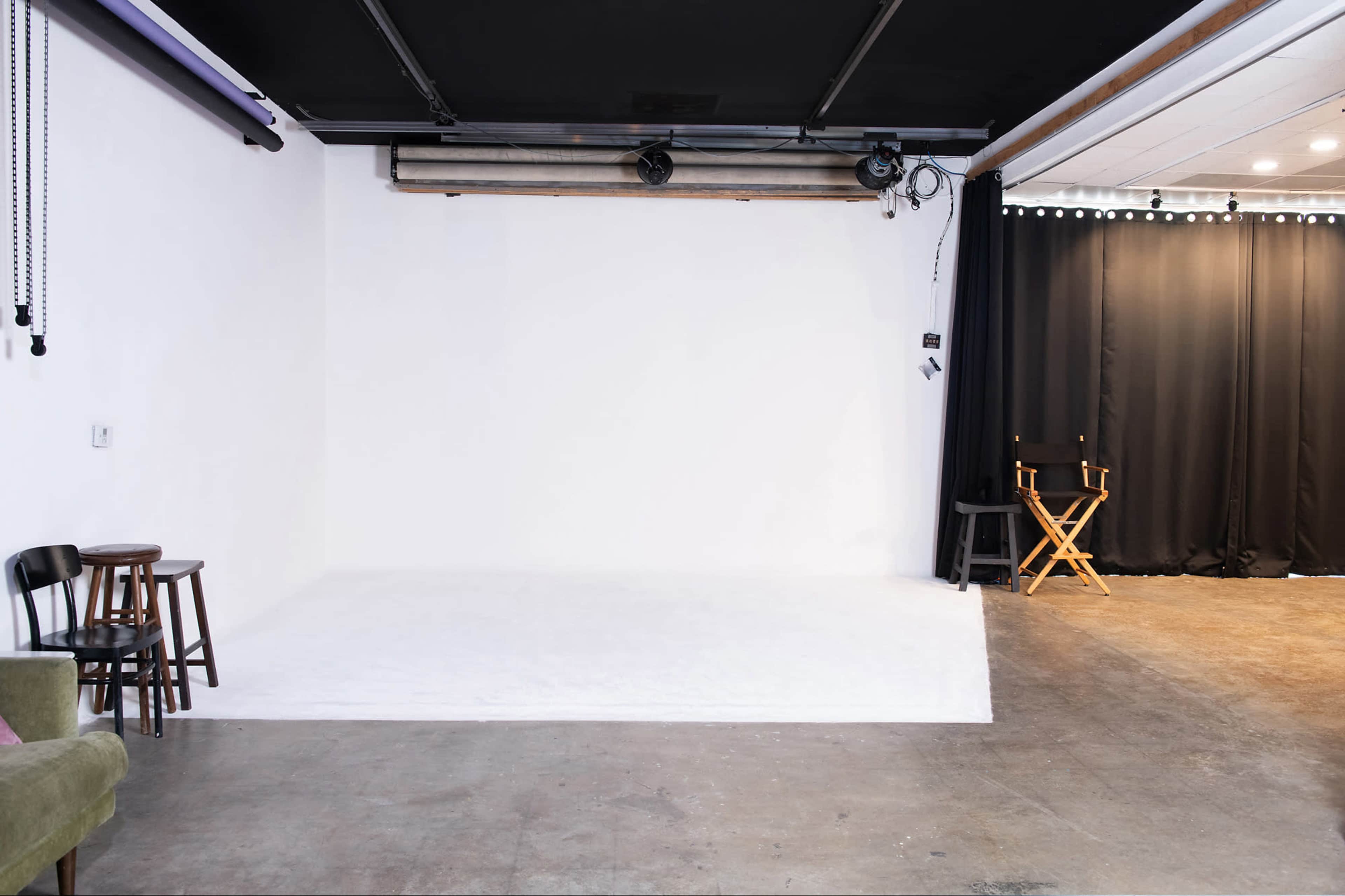 The image shows a photography studio featuring a white backdrop, a concrete floor, and a black curtain, with a few chairs positioned nearby.