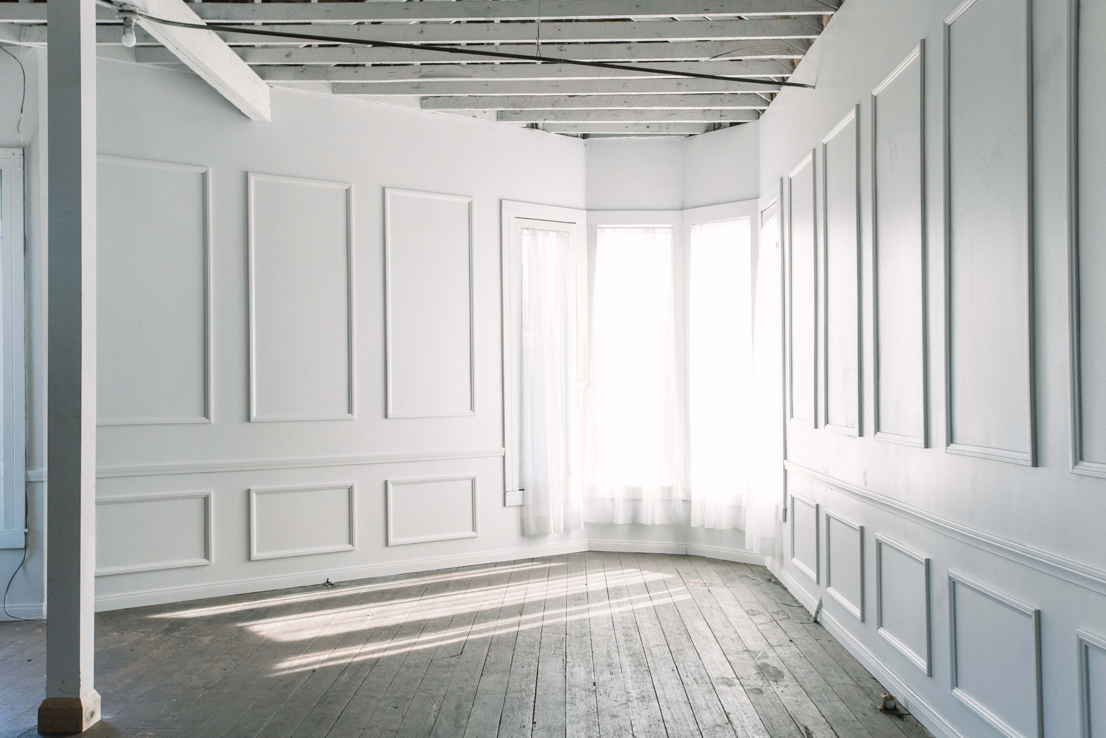A sunlit corner of a room features white walls with decorative paneling and bare wooden floors.