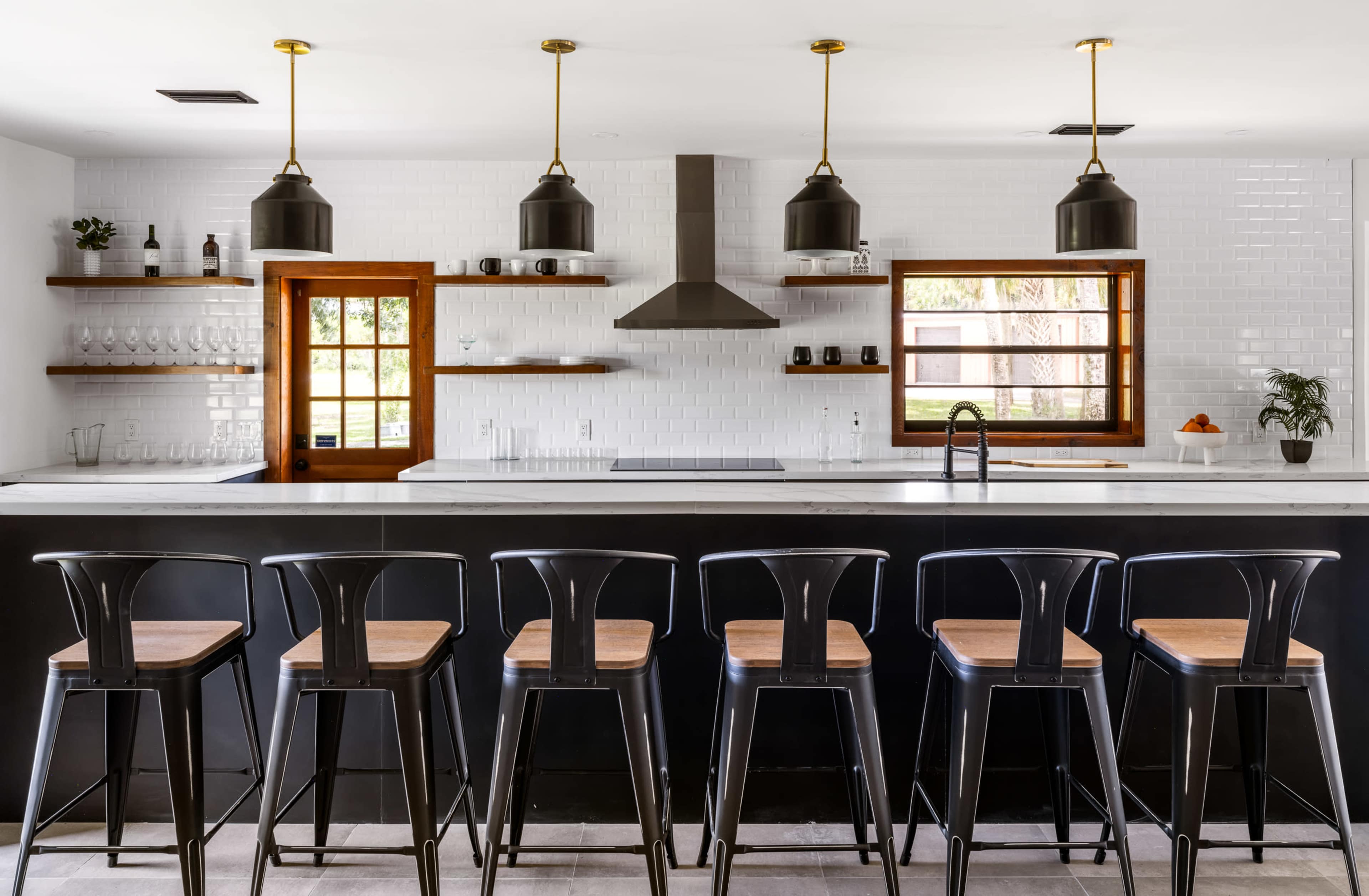 A modern kitchen features a long central island with black bar stools, overhead pendant lights, and white tiled walls with wooden shelves.