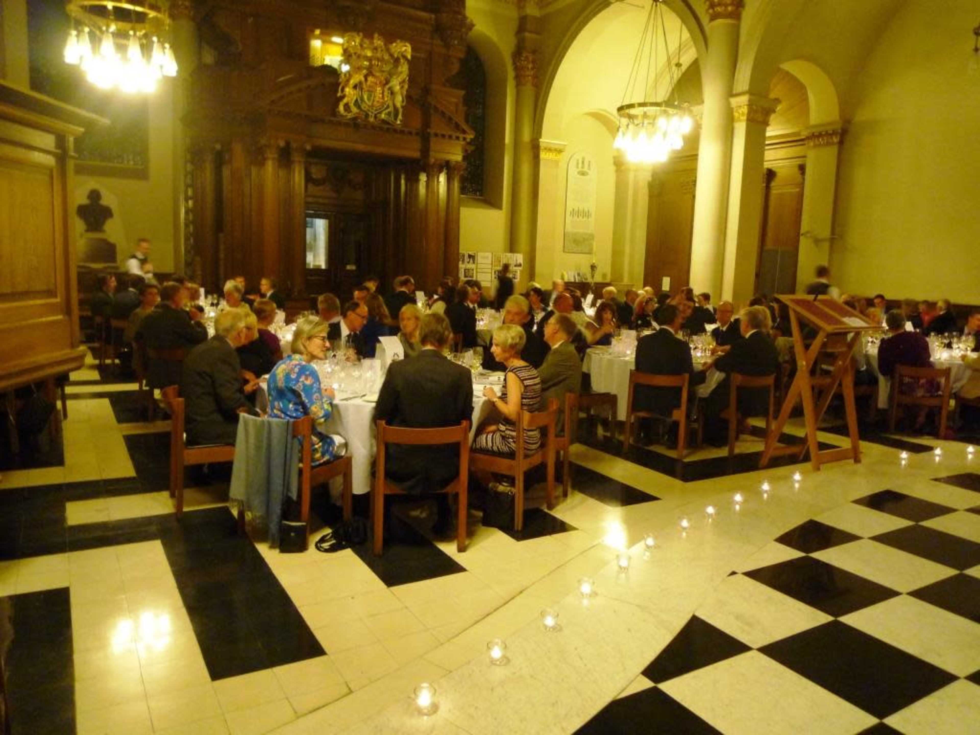 A formal dining event is taking place in a large hall, with numerous tables set for guests amidst elegant décor and candlelight.