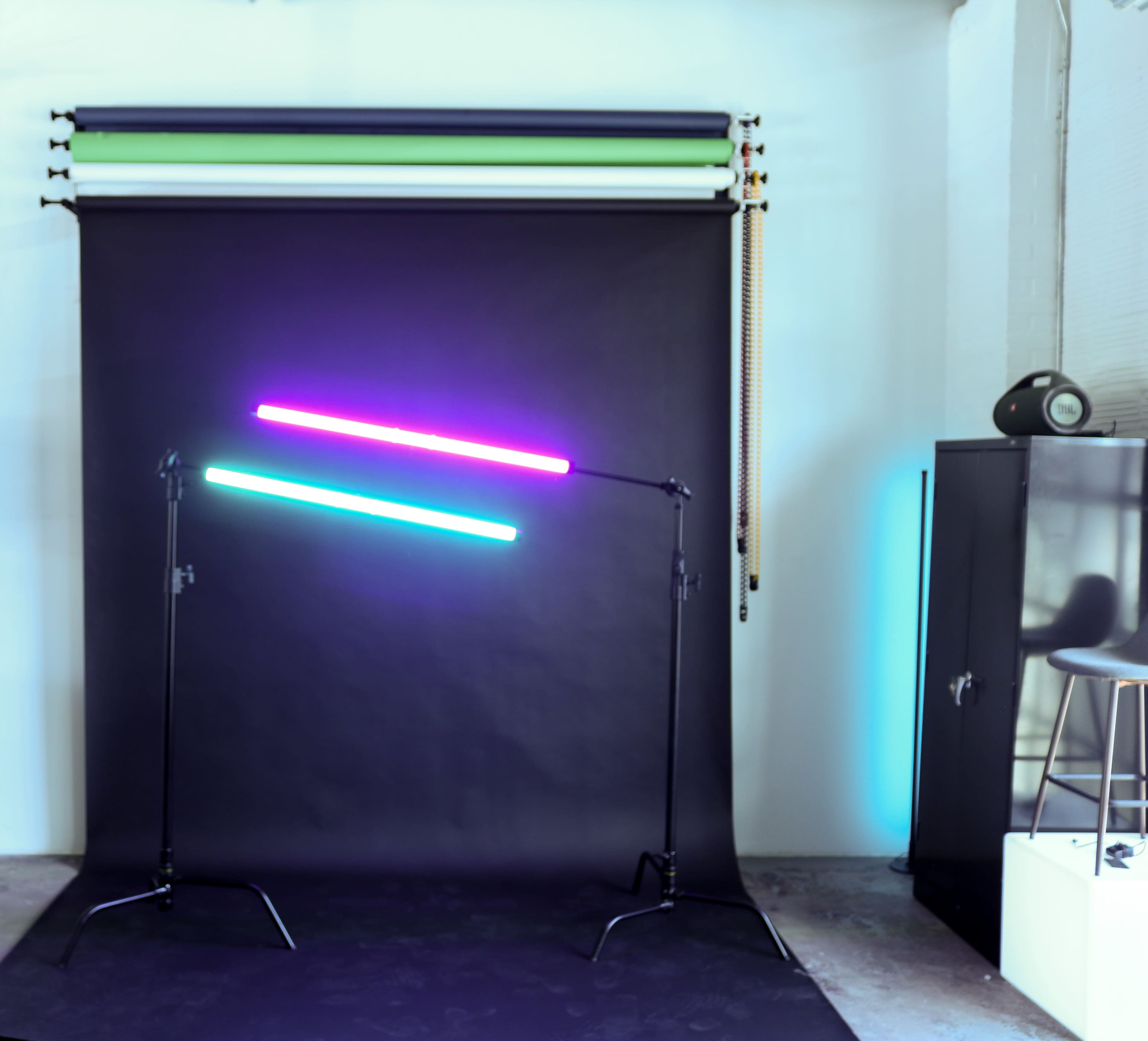 The image shows a photography studio setup with two colored light tubes positioned at angles against a dark backdrop, along with rolls of background paper hanging on the wall.