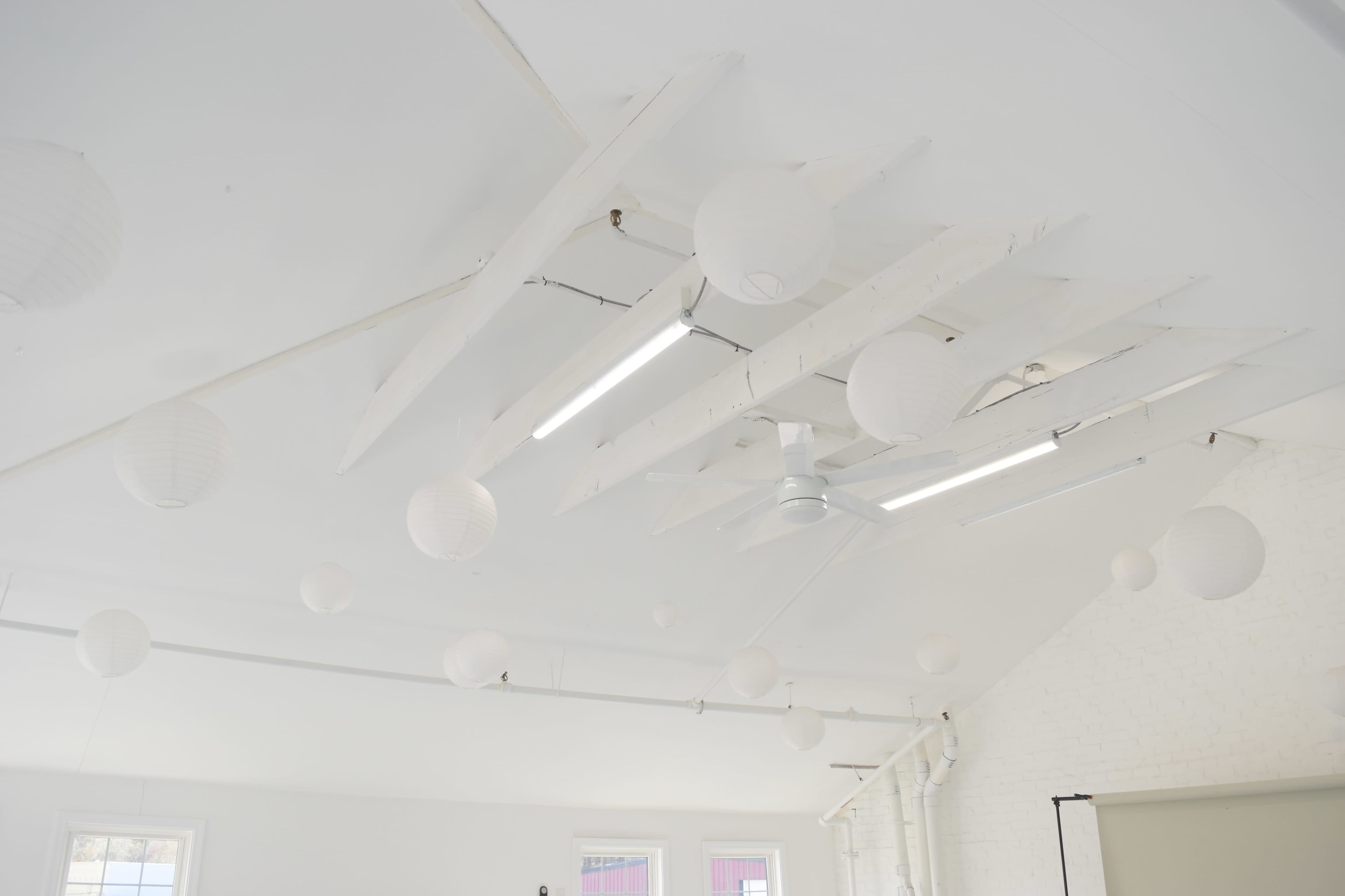 The image shows a bright, minimalist room with a white ceiling featuring wooden beams and round paper lanterns hanging down.