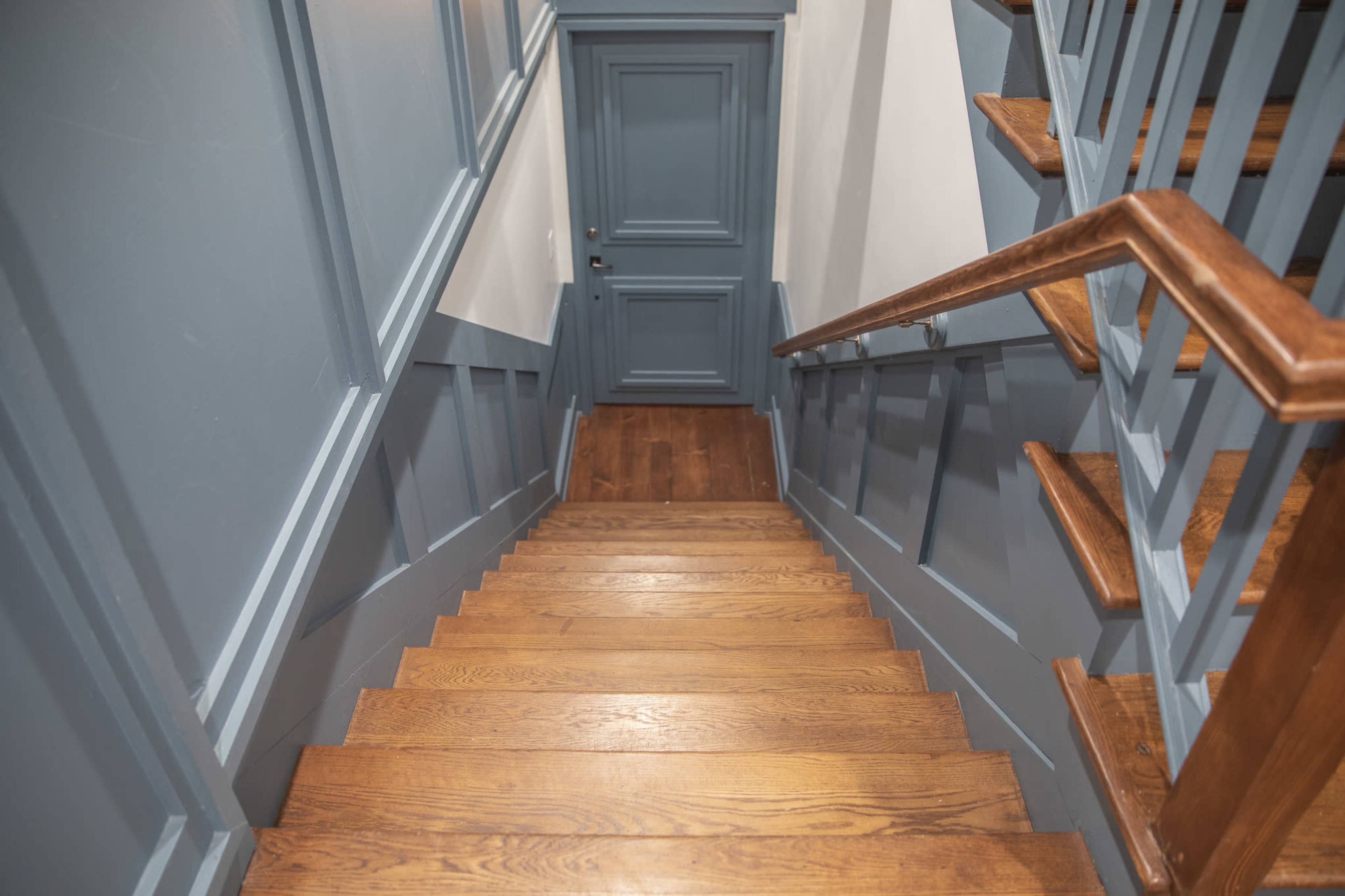 A wooden staircase leads down to a closed blue door at the bottom, framed by gray walls and wooden railings.