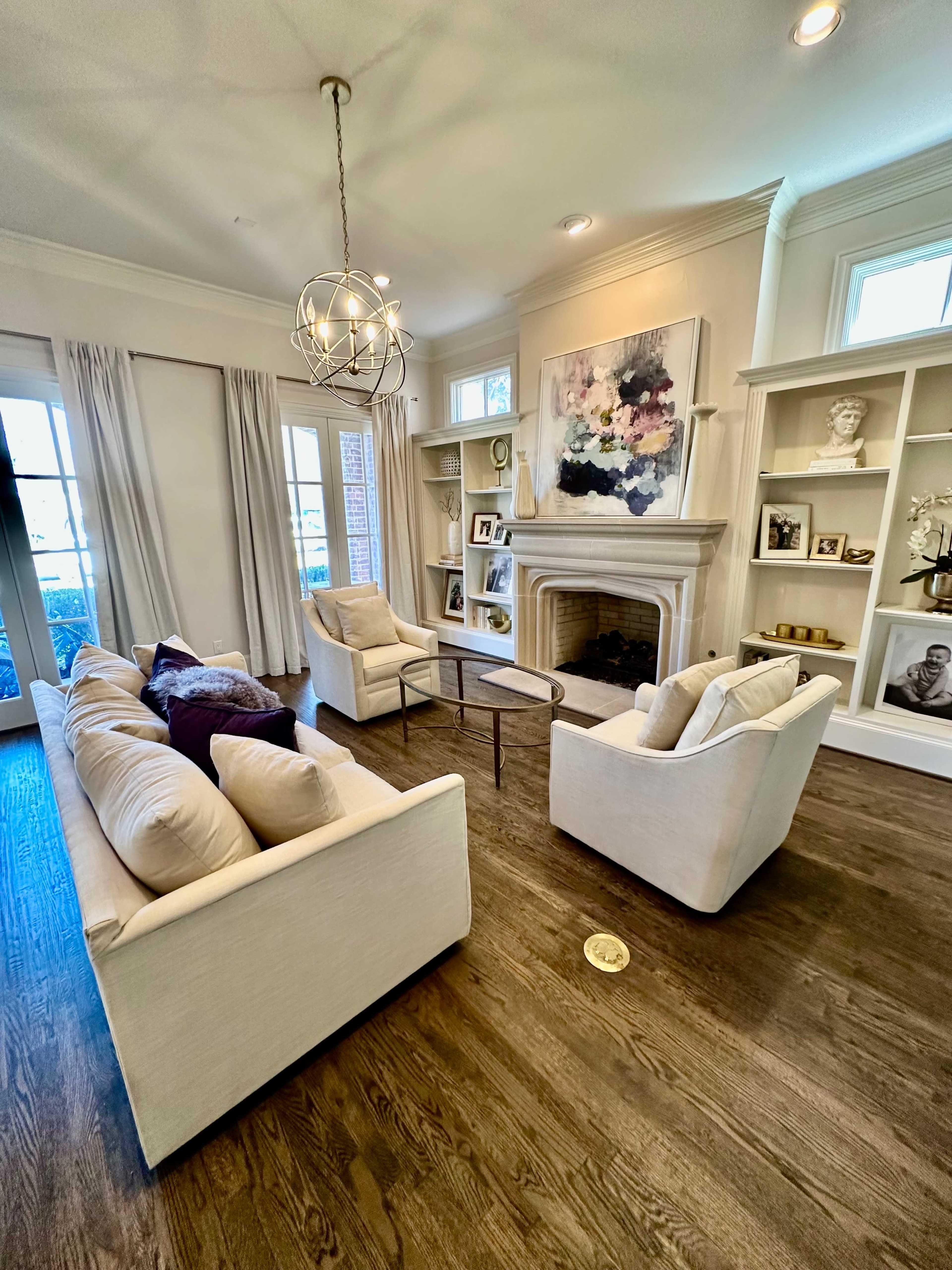 The image shows a well-lit living room with two white sofas facing a central coffee table and a fireplace, flanked by built-in shelves containing various decorative items and artwork.