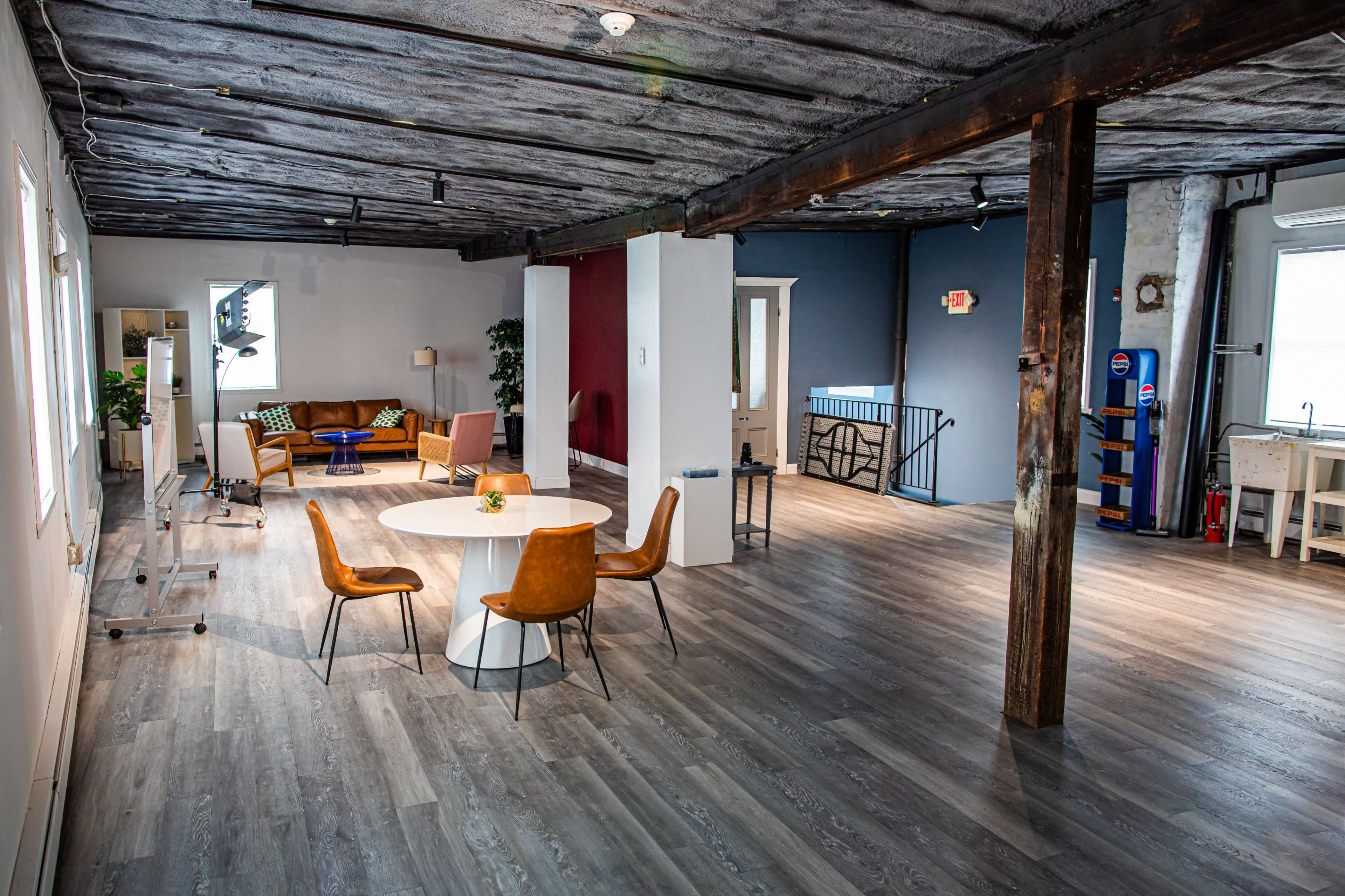 The image shows a spacious room with wooden floors, a circular table surrounded by four chairs, and a seating area with orange and brown furniture.