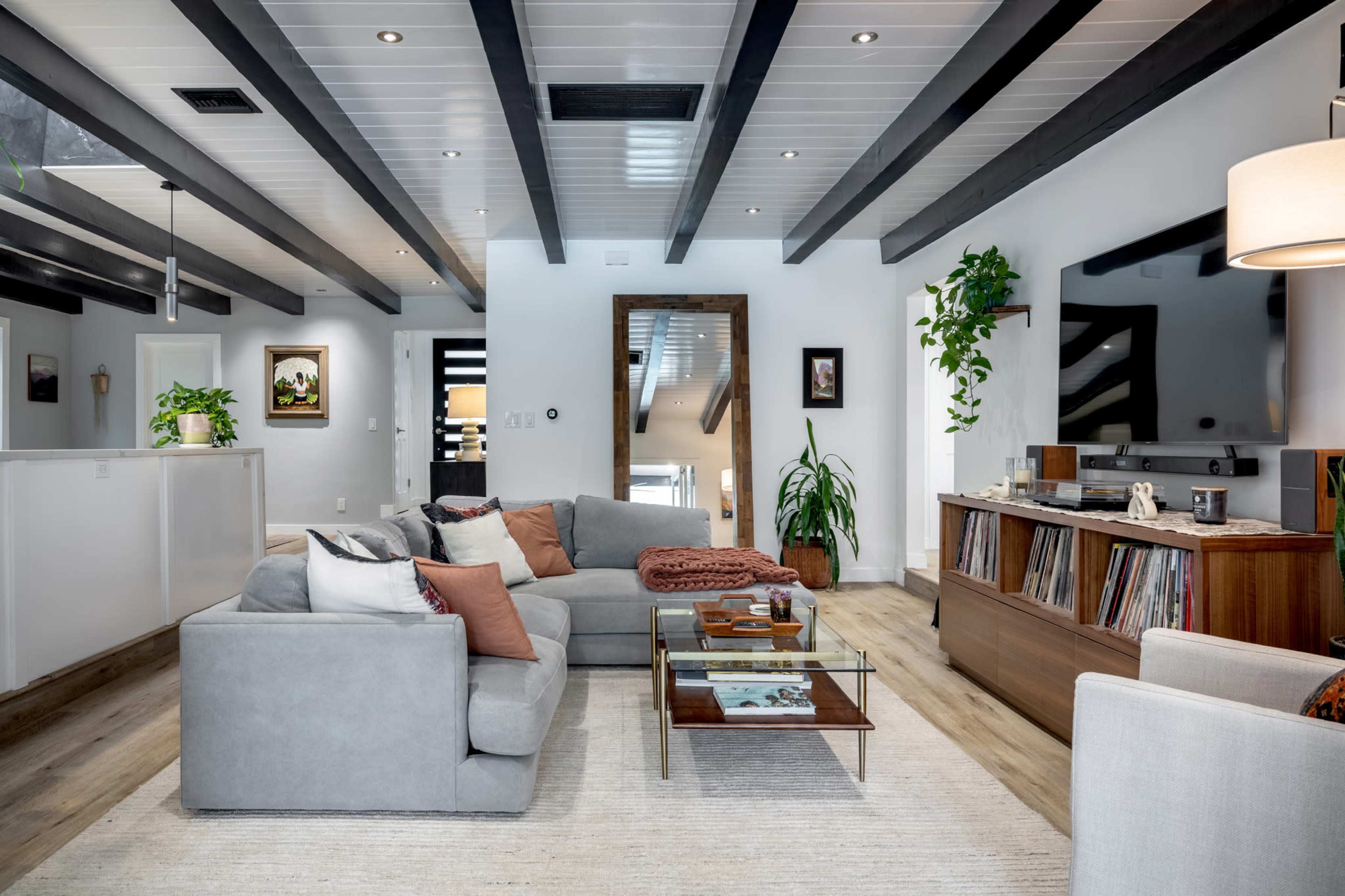The living room features a gray sectional sofa, a glass coffee table, wooden shelving with decor, and large windows with a view of greenery.