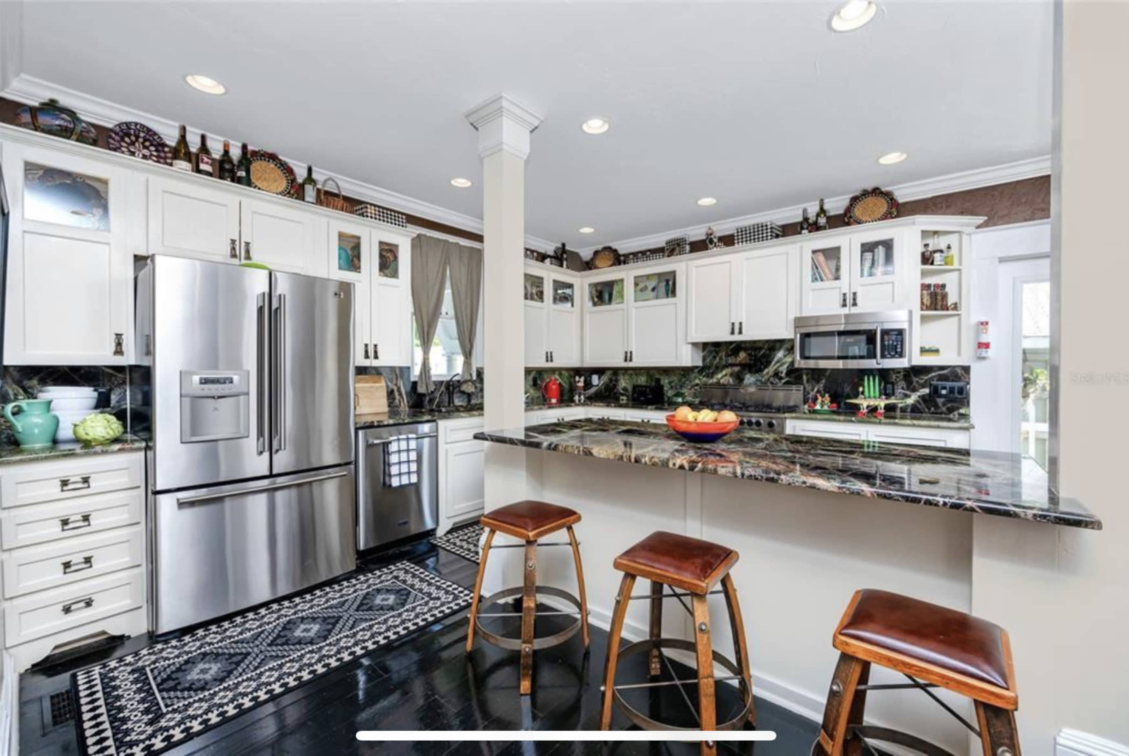 The image shows a modern kitchen featuring stainless steel appliances, granite countertops, and bar stools arranged around a small island.