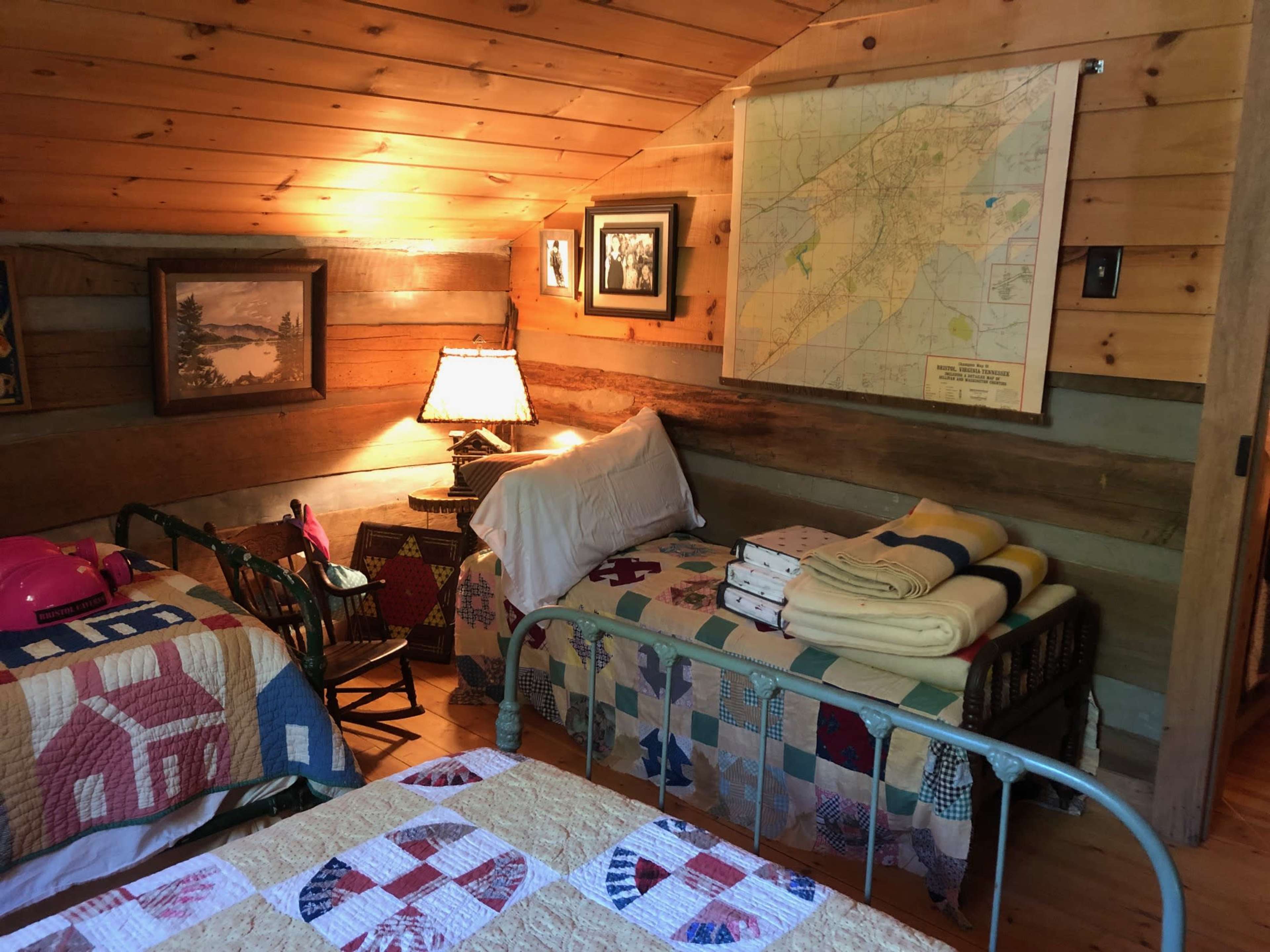A cozy attic bedroom with wooden walls, two beds covered with quilts, a lamp on a bedside table, and a map hanging on the wall.