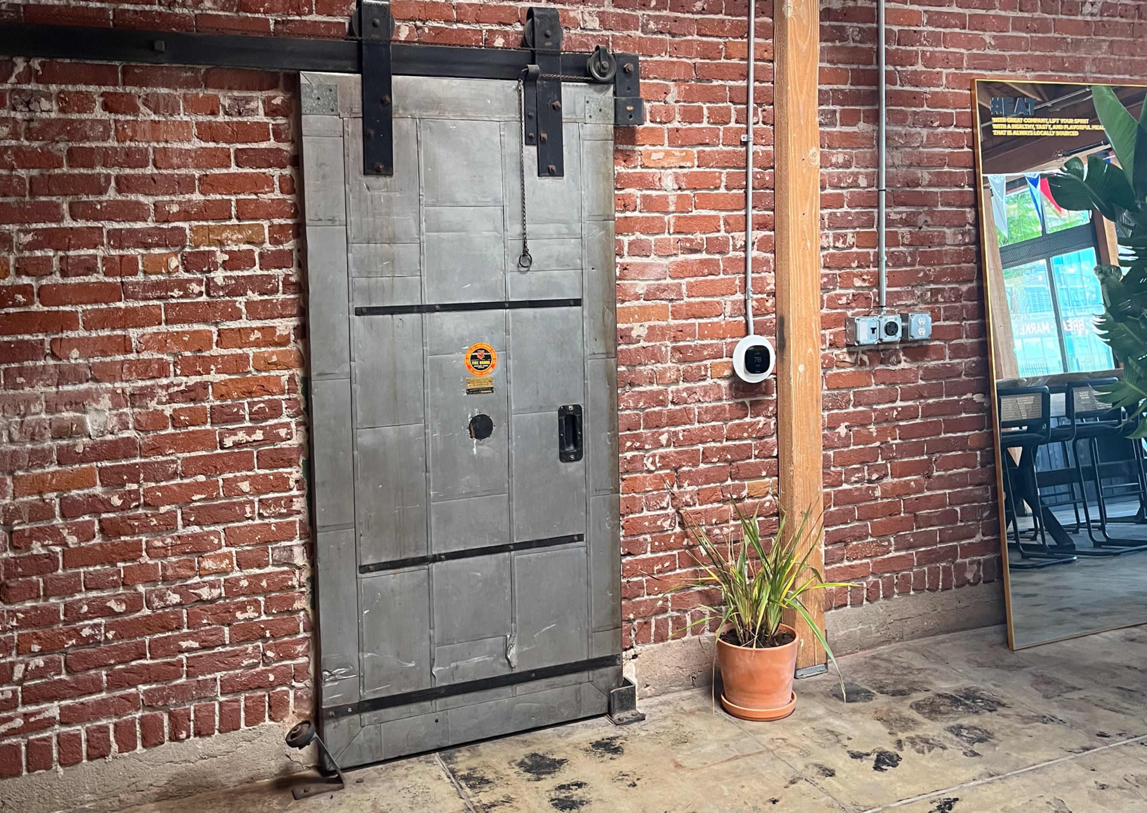 A metal sliding door mounted on a brick wall, with a potted plant and a large mirror nearby.