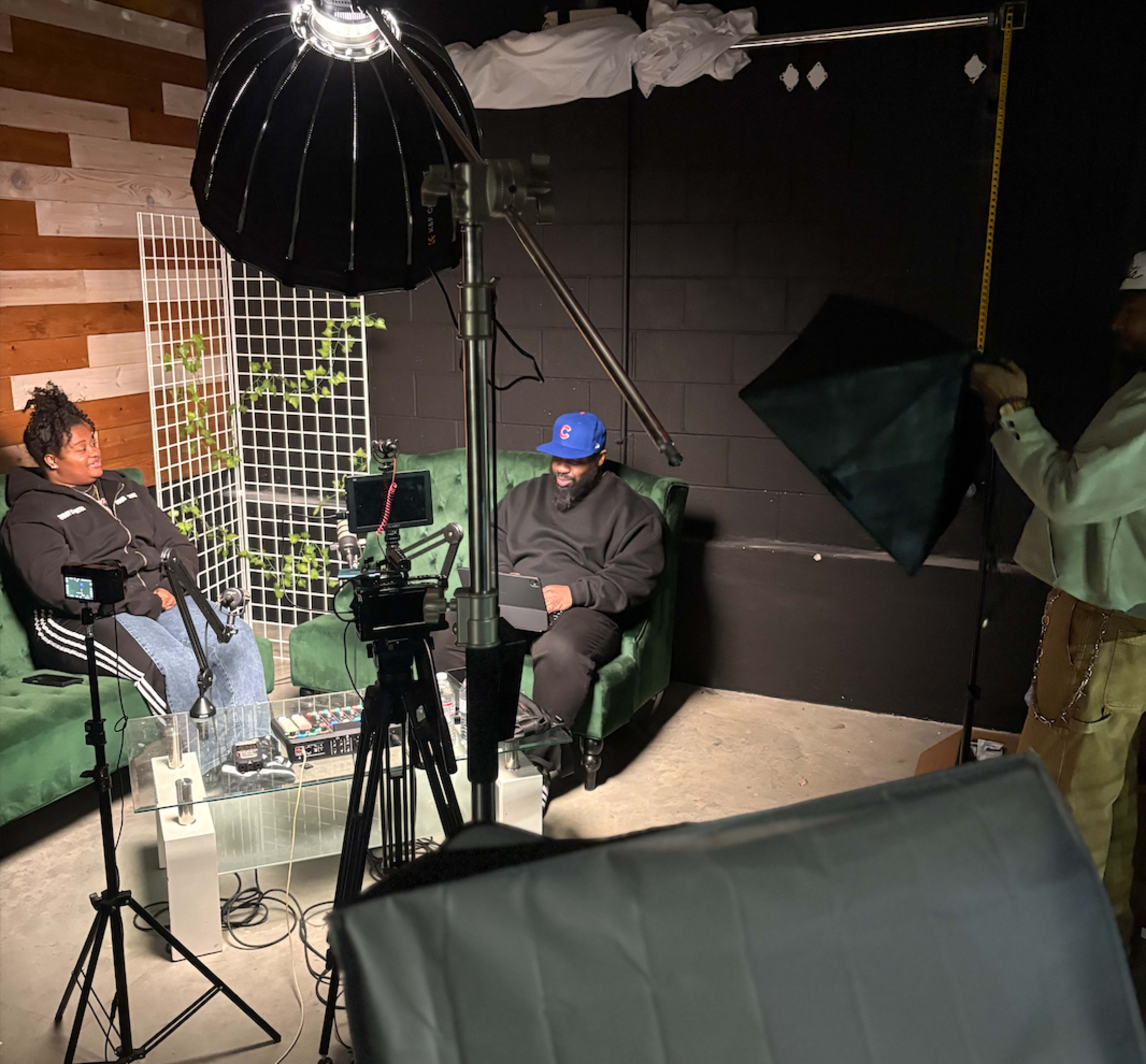 The image shows a studio setup with two people sitting on green couches, surrounded by camera equipment and lighting for an interview or recording session.