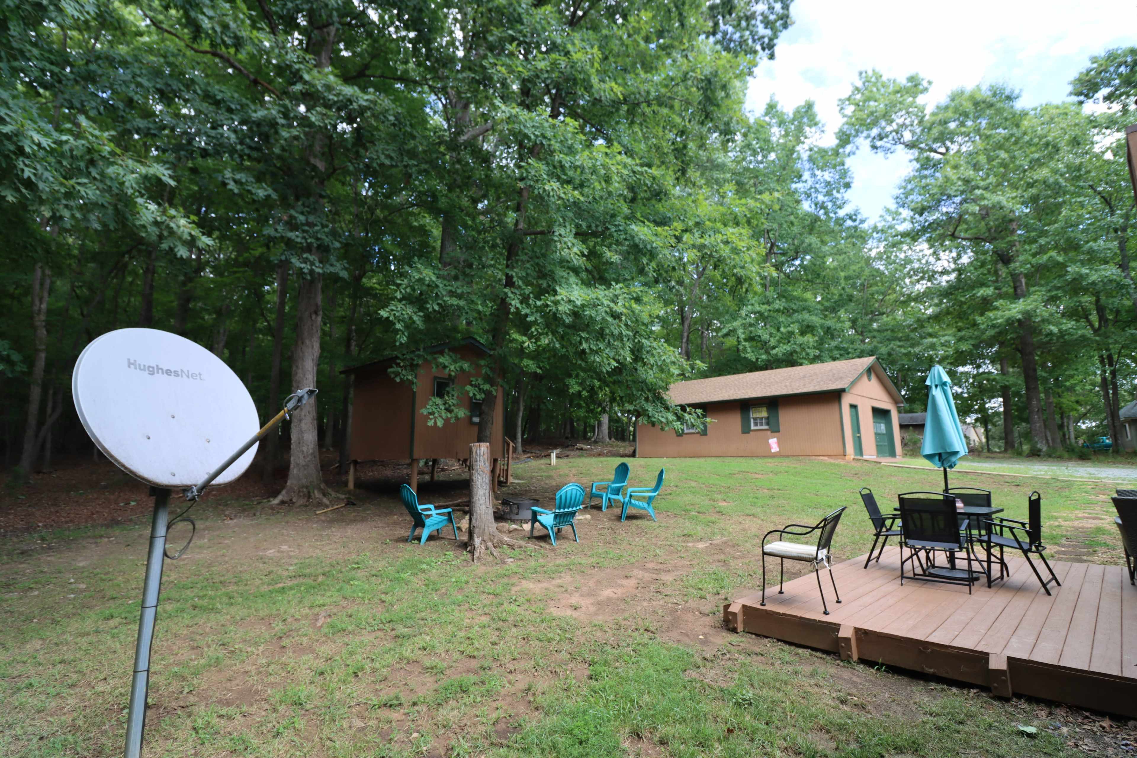 A wooded area featuring a brown building, a small structure, a satellite dish, and outdoor seating with a patio table and chairs.