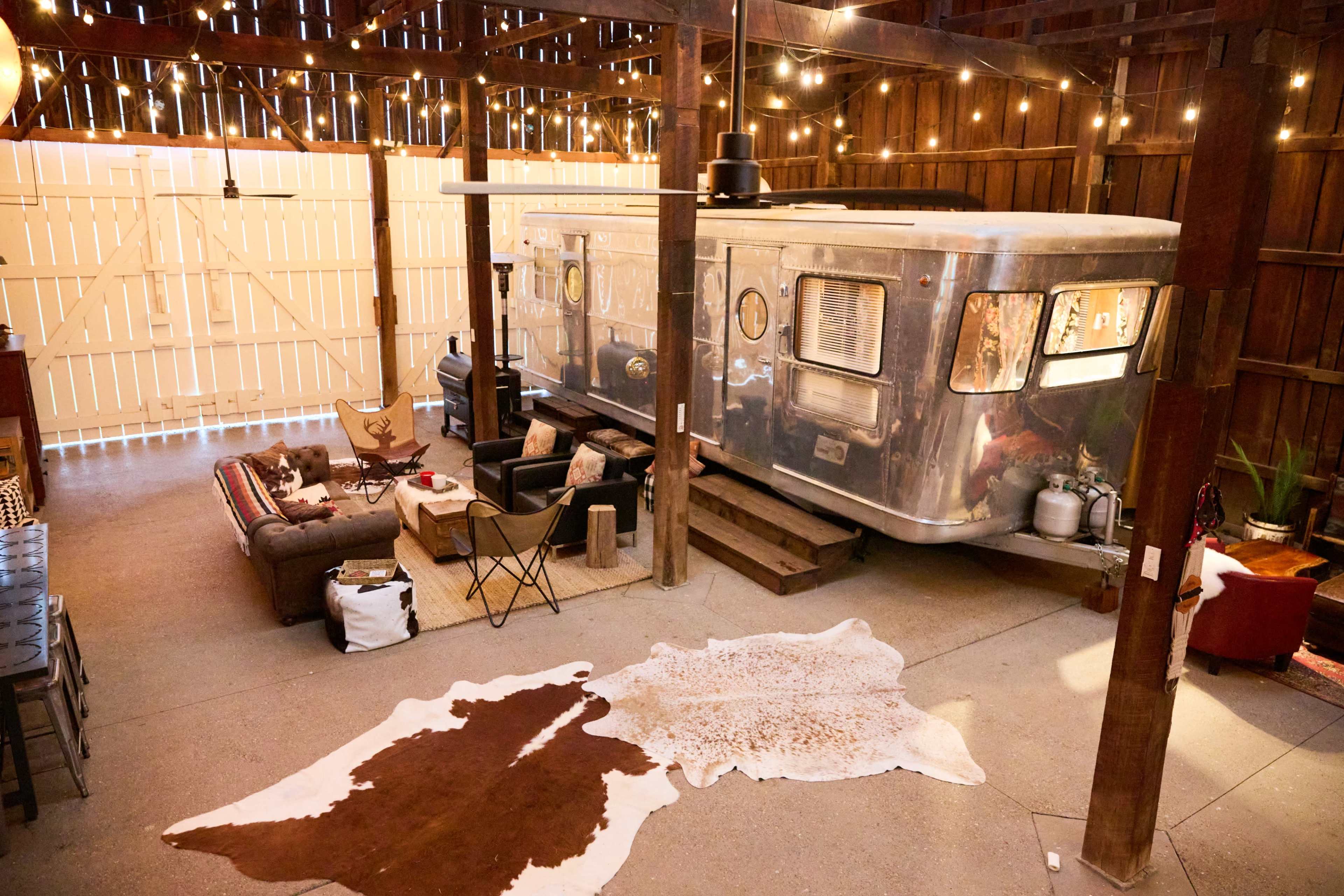 An agricultural barn interior features an airstream trailer surrounded by furniture, decorative rugs, and warm lighting.