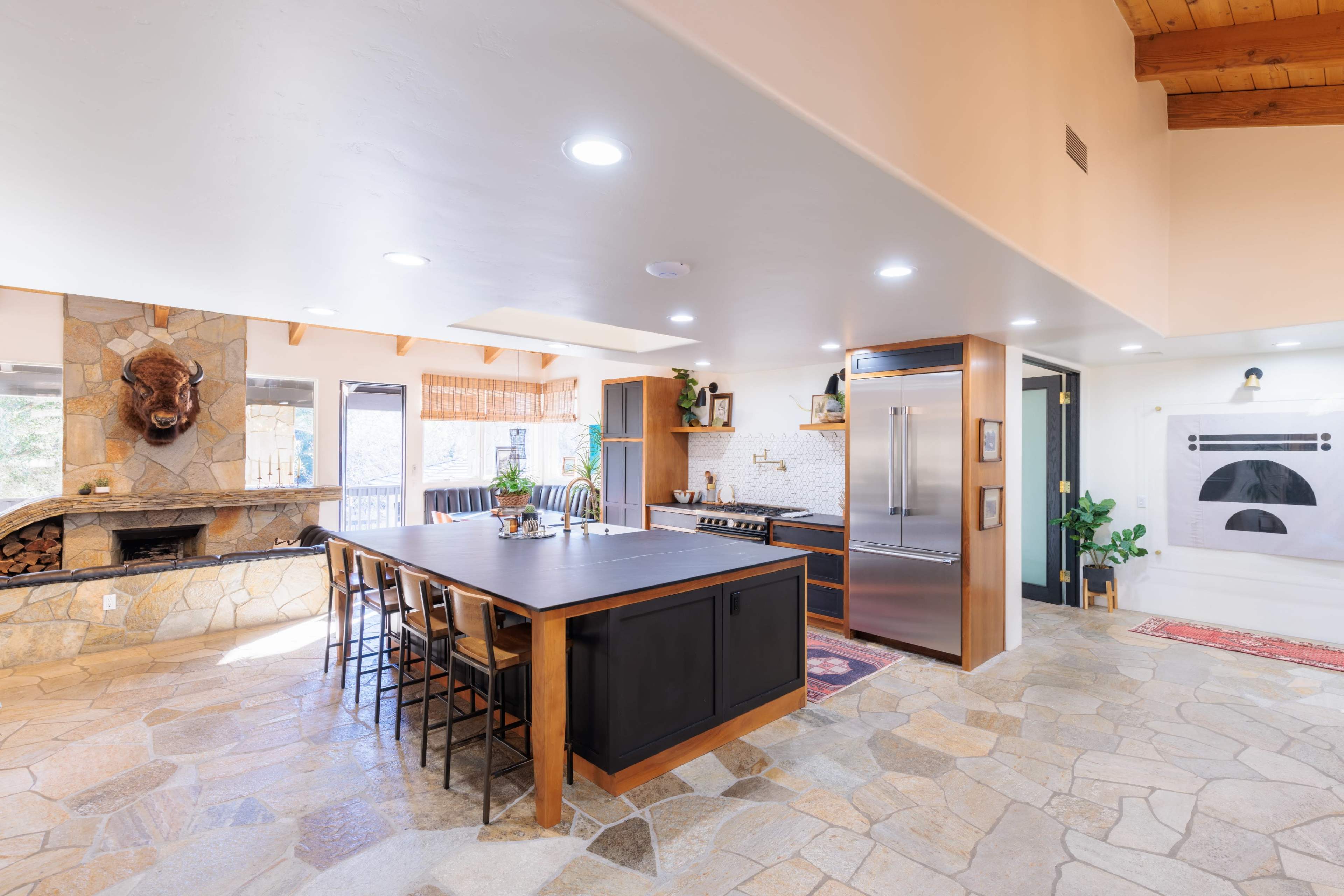 The image shows a modern kitchen with a large central island, stainless steel appliances, a stone fireplace, and a combination of wooden and stone flooring.
