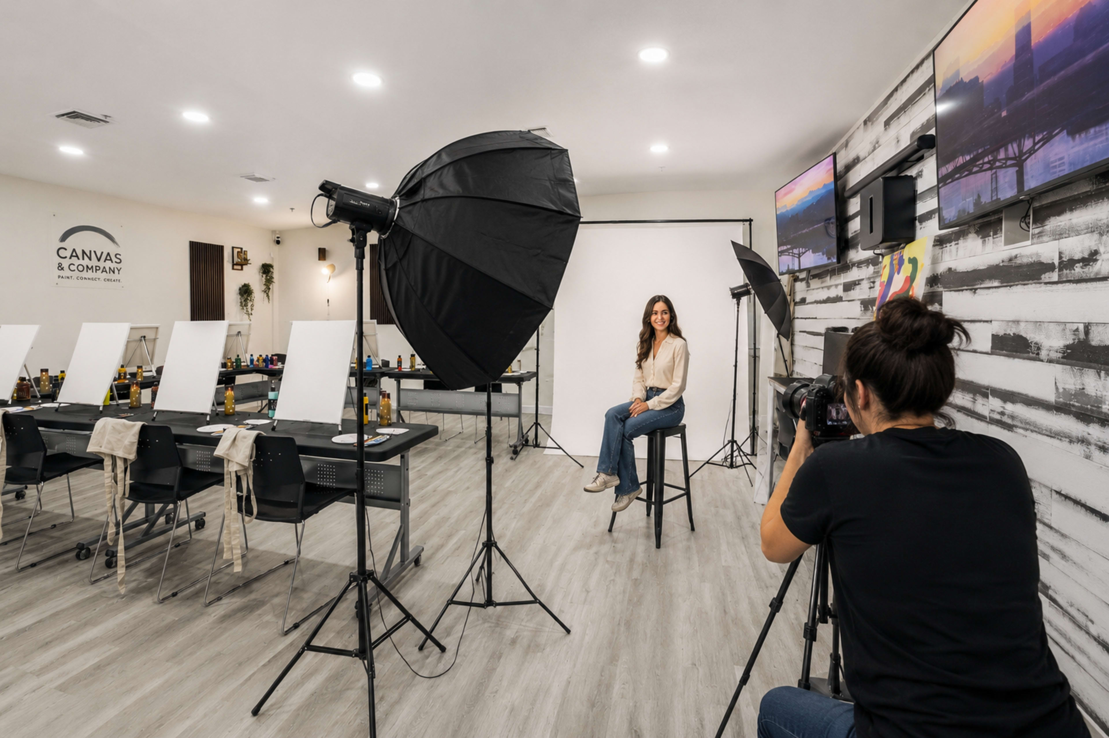 A photographer captures a portrait of a model sitting on a stool in a well-lit studio featuring multiple canvases and lighting equipment.