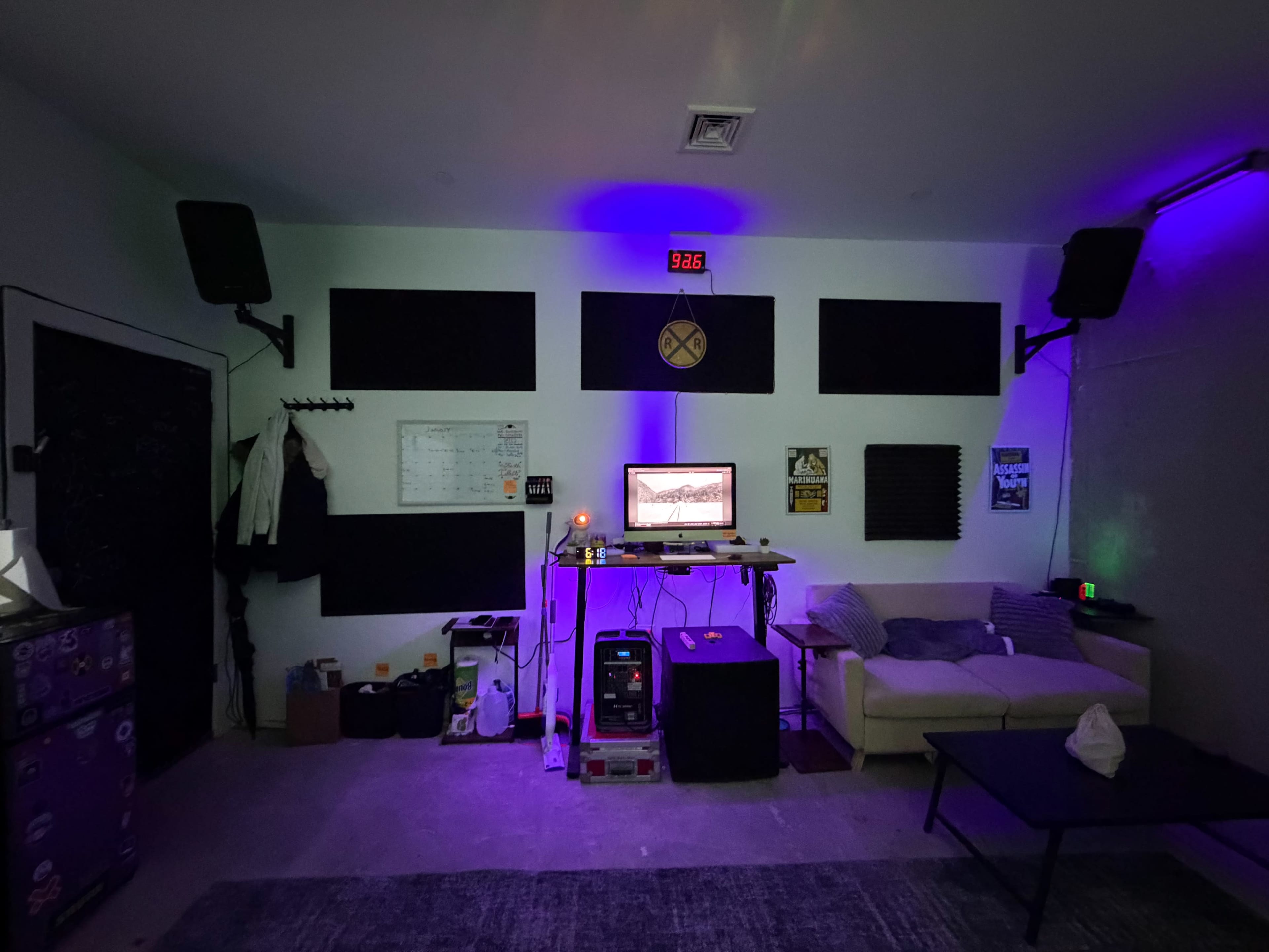 The image shows a dimly lit room with a desk, computer setup, acoustic panels on the walls, and a couch.