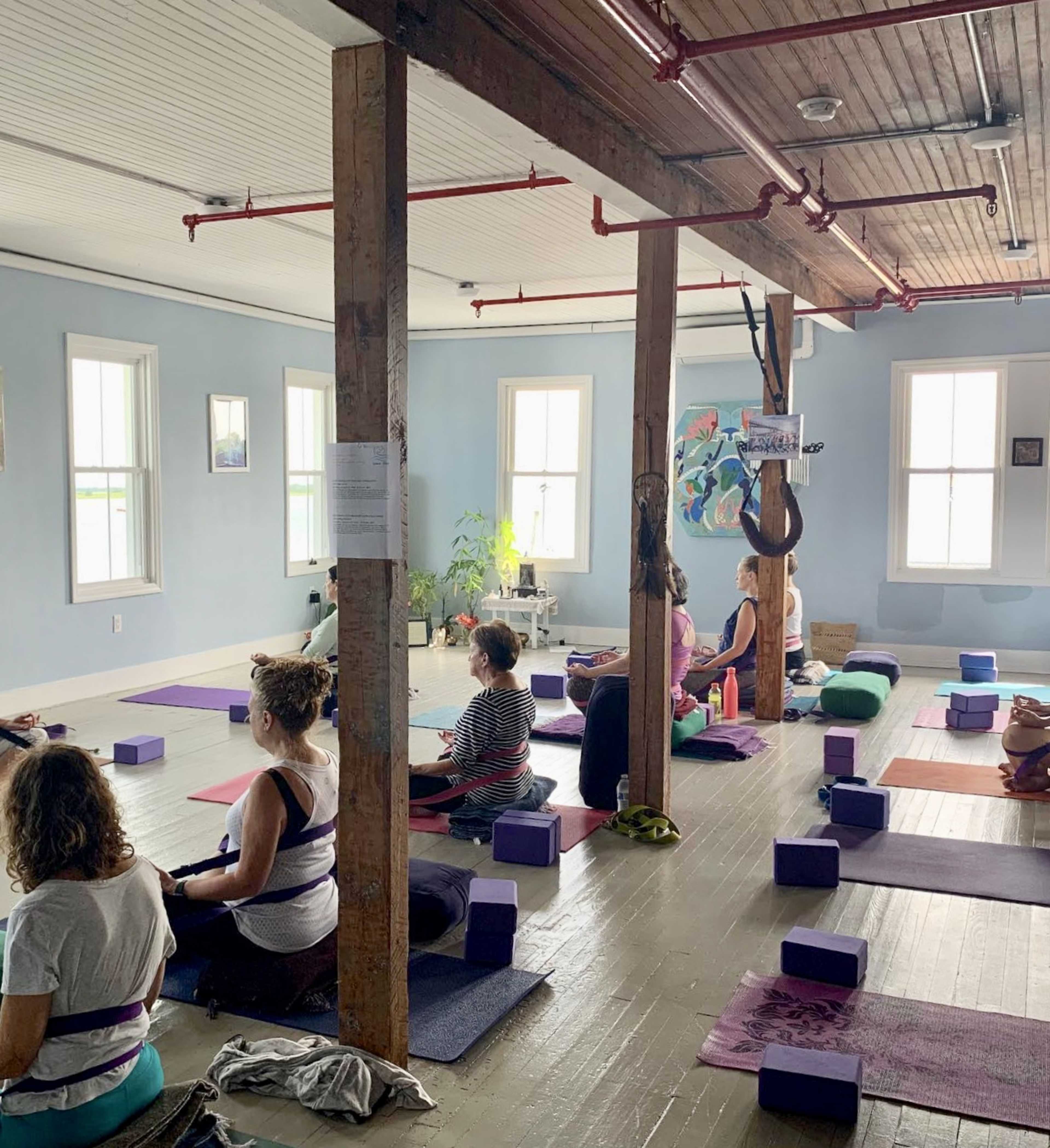 A group of people practice yoga in a brightly lit studio with wooden beams and various yoga props scattered on the floor.