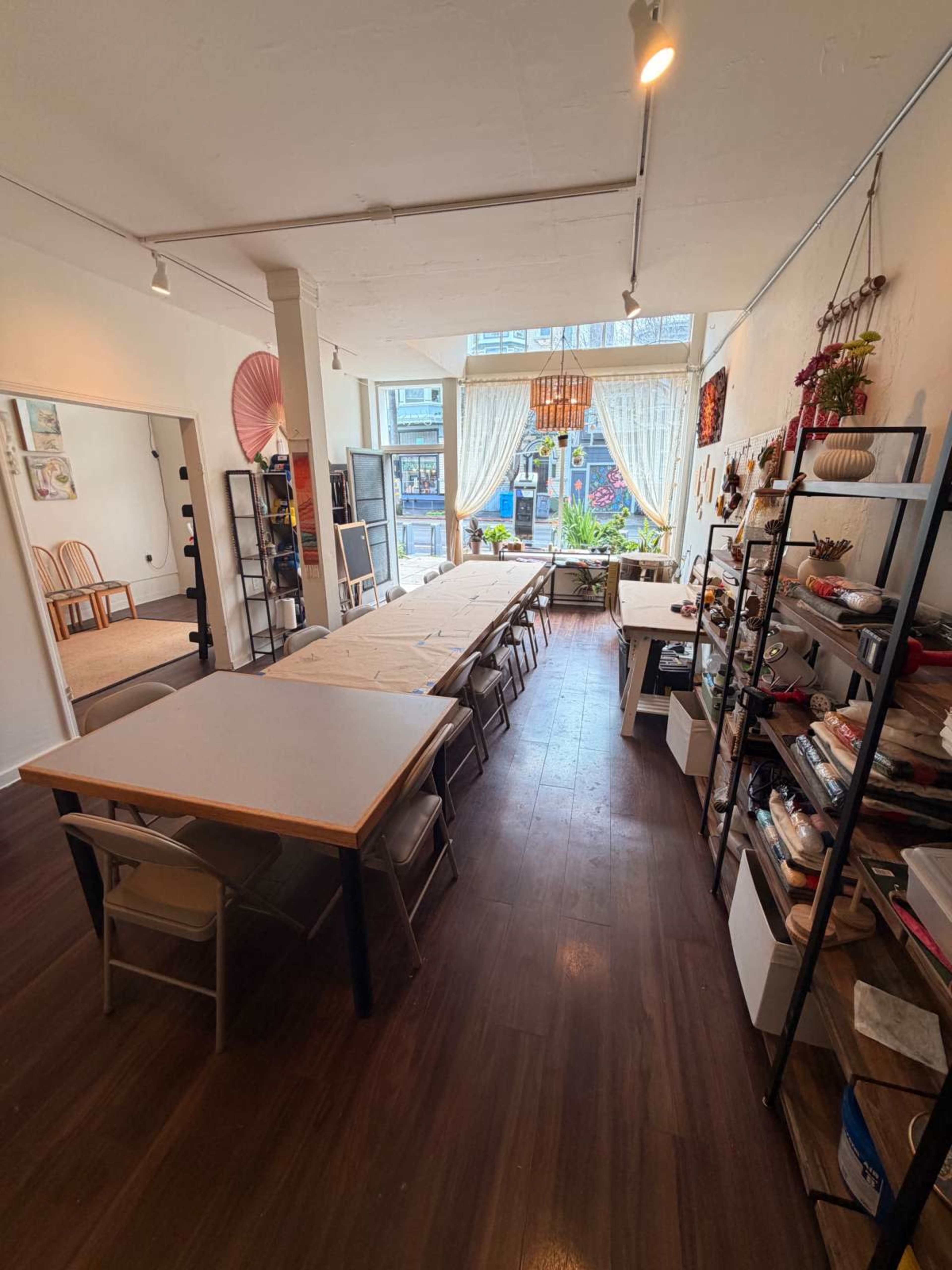 The image shows a spacious room set up for a gathering, featuring a long table with chairs, shelves filled with various items, and large windows allowing natural light to fill the space.